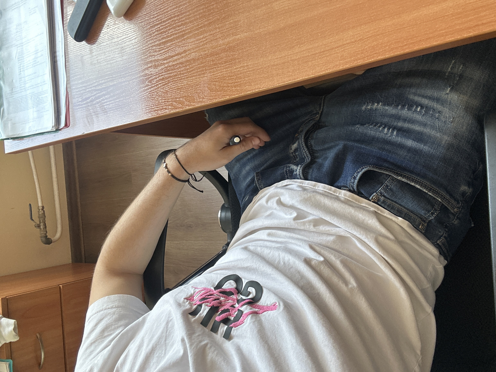 A medium shot from an elevated angle depicts a person seated at a wooden desk. The individual is wearing blue jeans and a white t-shirt with a distinctive logo featuring black numbers and a pink embellishment. The person's left hand rests on their lap, holding a dark object. The desk is covered with documents and office supplies. A cabinet is visible in the lower-left corner. Based on the presence of Cyrillic characters on the documents, the location may be within Bulgaria, and potentially in Plovdiv.