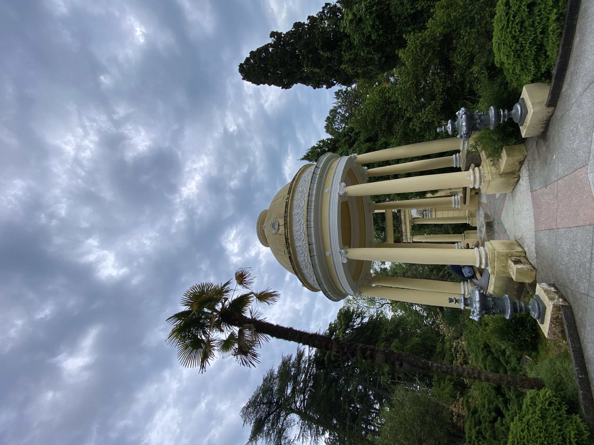 The image shows a gazebo in a park, with a cloudy sky above. The gazebo has a dome roof supported by columns. A palm tree stands near the gazebo. The location is likely Sochi, Russia.