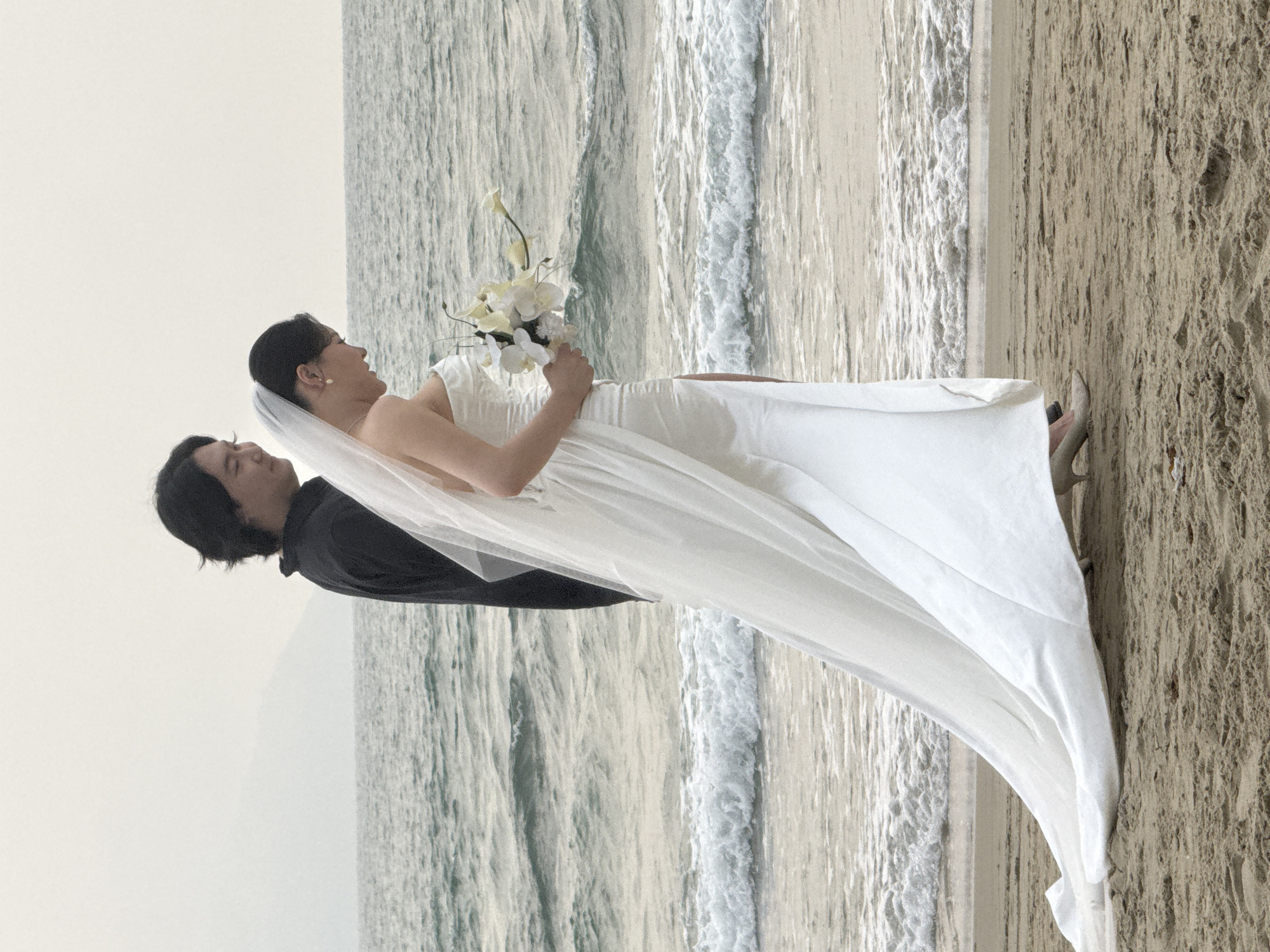 A bride and groom pose for a photo on a beach. The bride wears a white dress, veil, and holds a bouquet of white flowers. The groom wears a black shirt. They are standing near the water's edge. The background consists of the ocean and a sandy beach.
It is impossible to pinpoint the specific beach location within Đà Nẵng, Vietnam, without additional information.