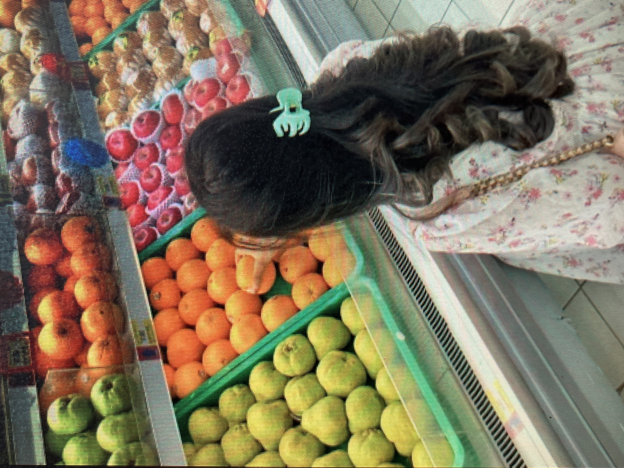 The image shows a person from behind, with dark curly hair, standing in front of a refrigerated display of fruits in a supermarket. The fruits are arranged in green crates and appear to be apples, oranges, and possibly pears. The person is wearing a floral patterned dress and a green hair clip. The environment suggests a well-lit supermarket with a wide selection of fresh produce. There is no specific information in the image that would allow for the identification of the city of Sejahtera, Indonesia.