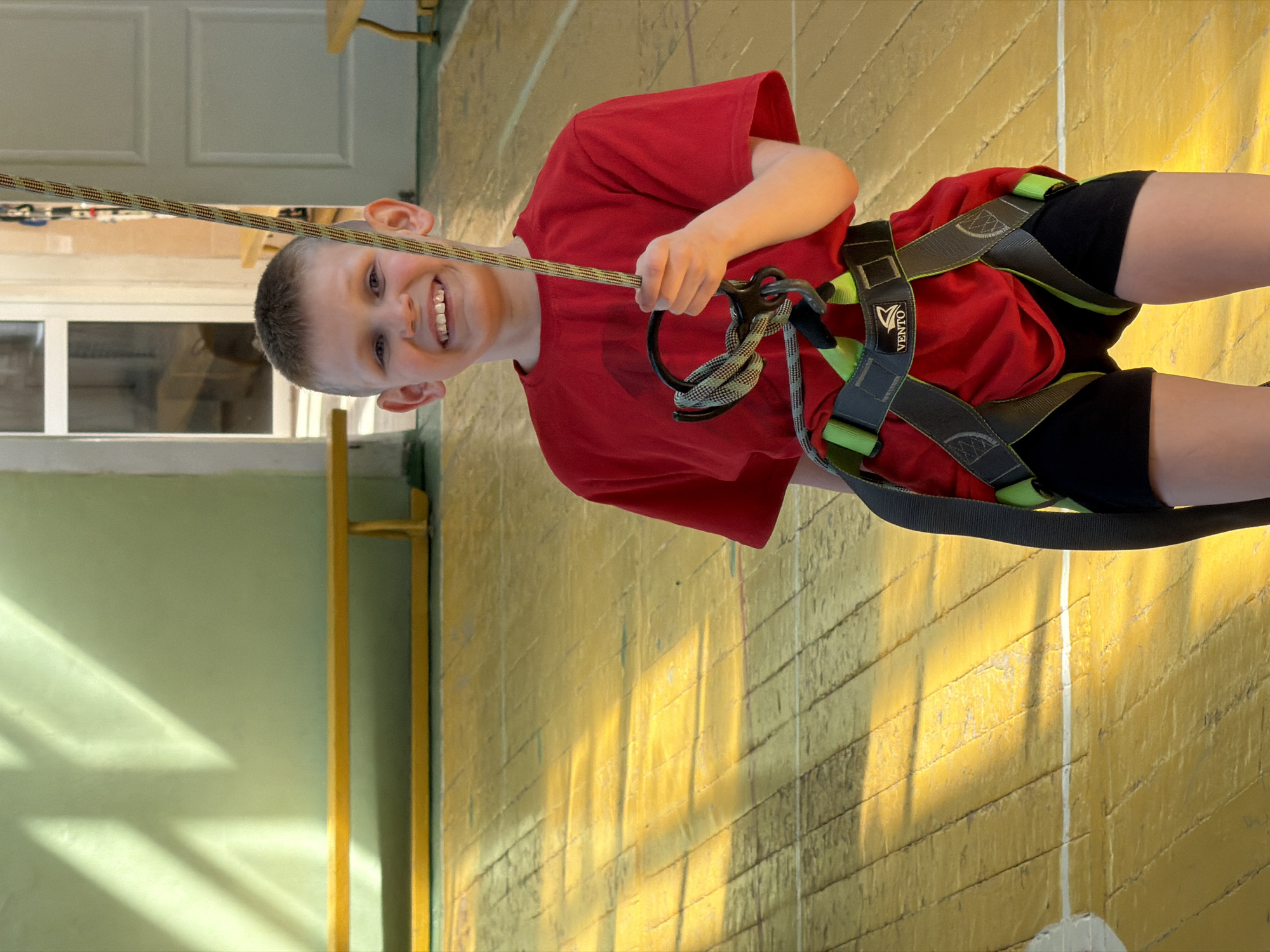 A young boy in a red shirt and black shorts is wearing a climbing harness indoors and holding a rope. The harness is gray and has green accents. The boy is smiling and looking at the camera. He is standing against a brick wall painted yellow, with a white stripe running vertically. The background includes a light green wall and a white door with a window, suggesting an indoor space like a gym or recreational facility. While the image does not inherently indicate the location in Izhevsk, Russia, there's no information within the image to confirm or deny this.