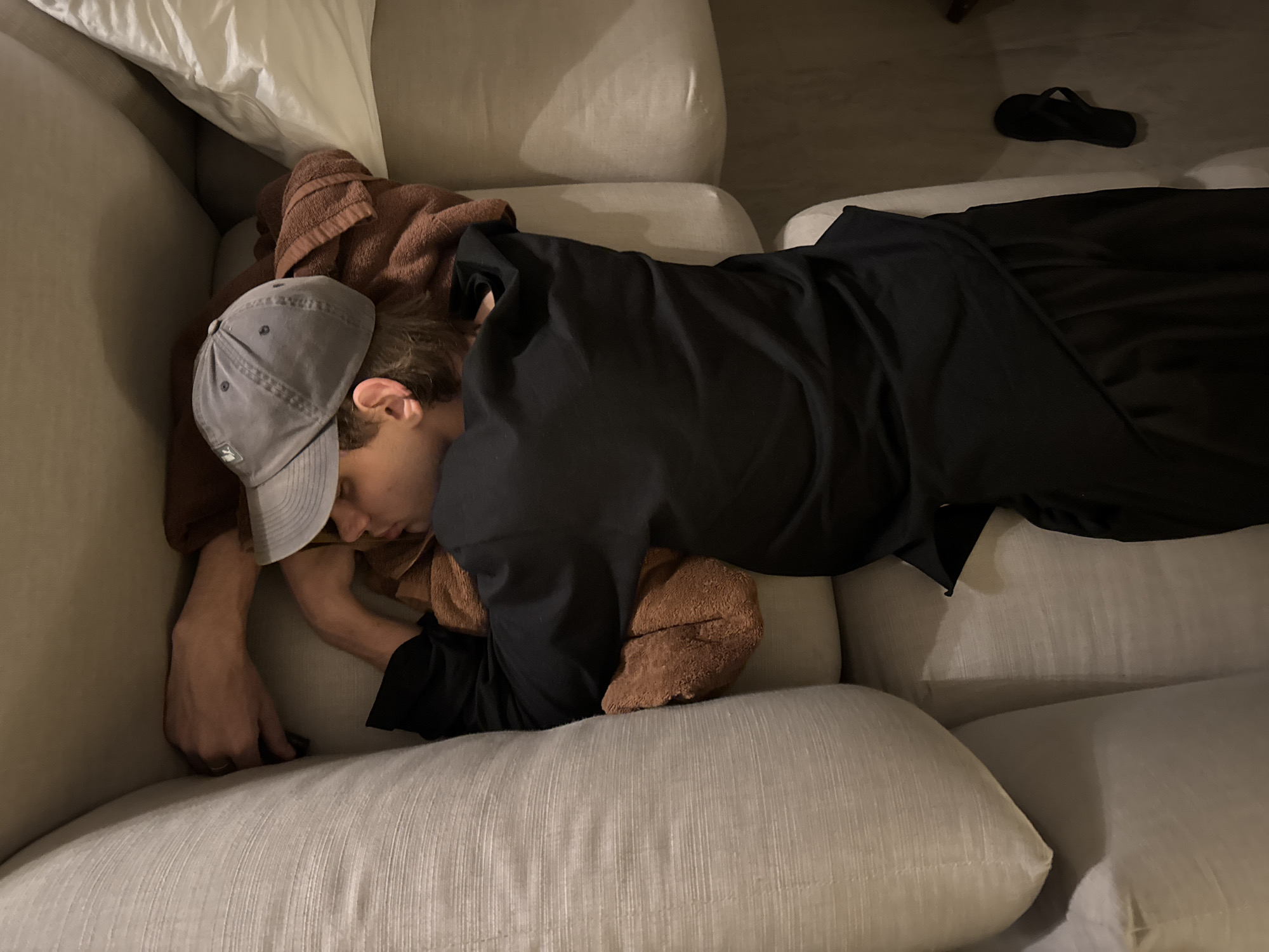 A person wearing a gray baseball cap and black clothing is asleep on a light-colored couch. A brown towel is draped over the person's head and shoulders. A white pillow is positioned behind the person's head. A pair of black flip-flops are on the floor in the background. The photo was taken indoors in Kuta Selatan, Indonesia.
