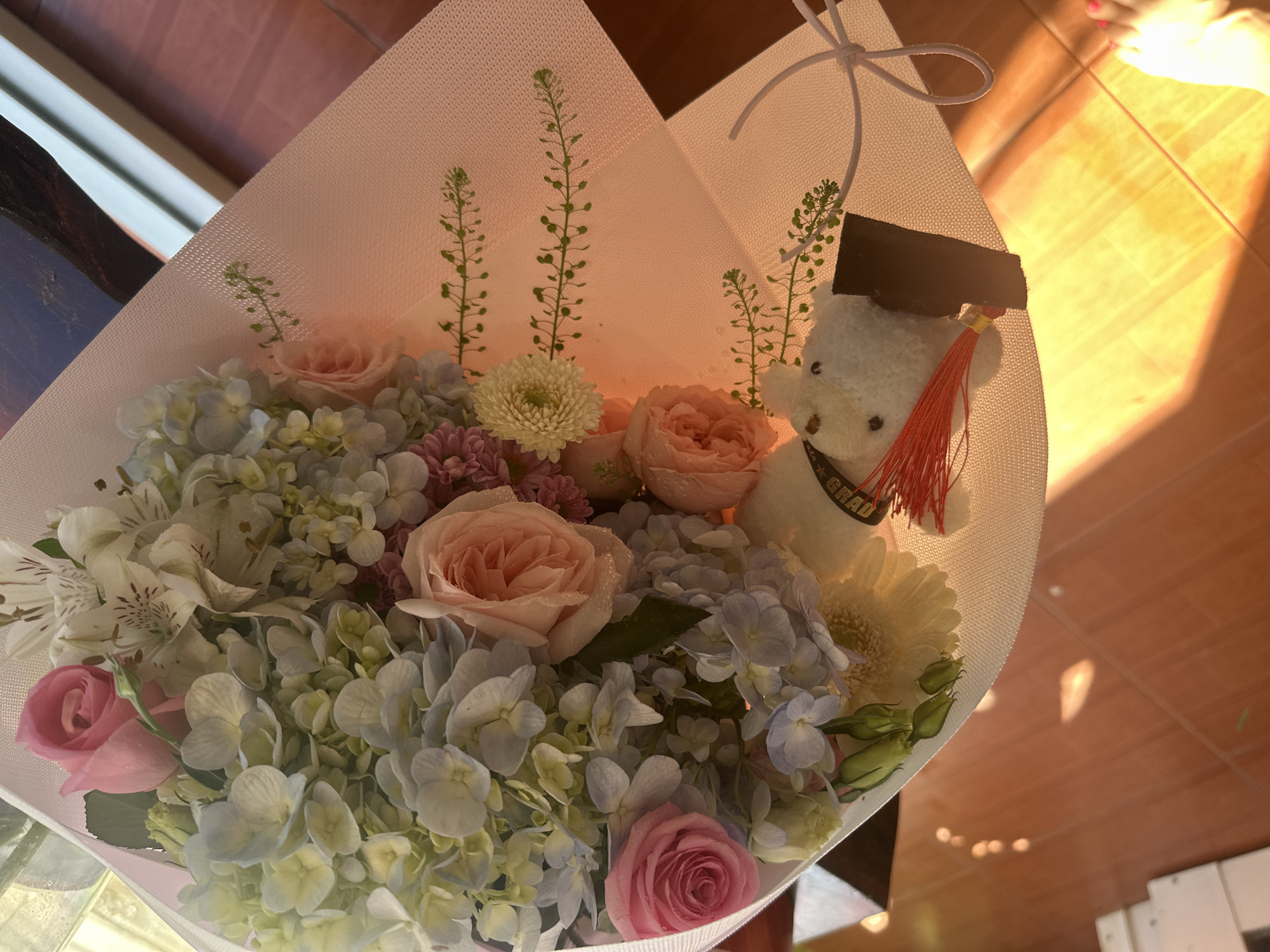 A high-angle shot of a flower bouquet in a white wrapper with a white ribbon tie, presented on a brown surface, likely a table. The bouquet features a mix of flowers including pink roses, light blue hydrangeas, white alstroemeria, and green foliage. In the center of the bouquet is a small, white teddy bear wearing a graduation cap with a red tassel and a sash that reads "GRAD". The bouquet is illuminated by natural light. The location is implied to be Bac Nha Trang Ward, Vietnam.