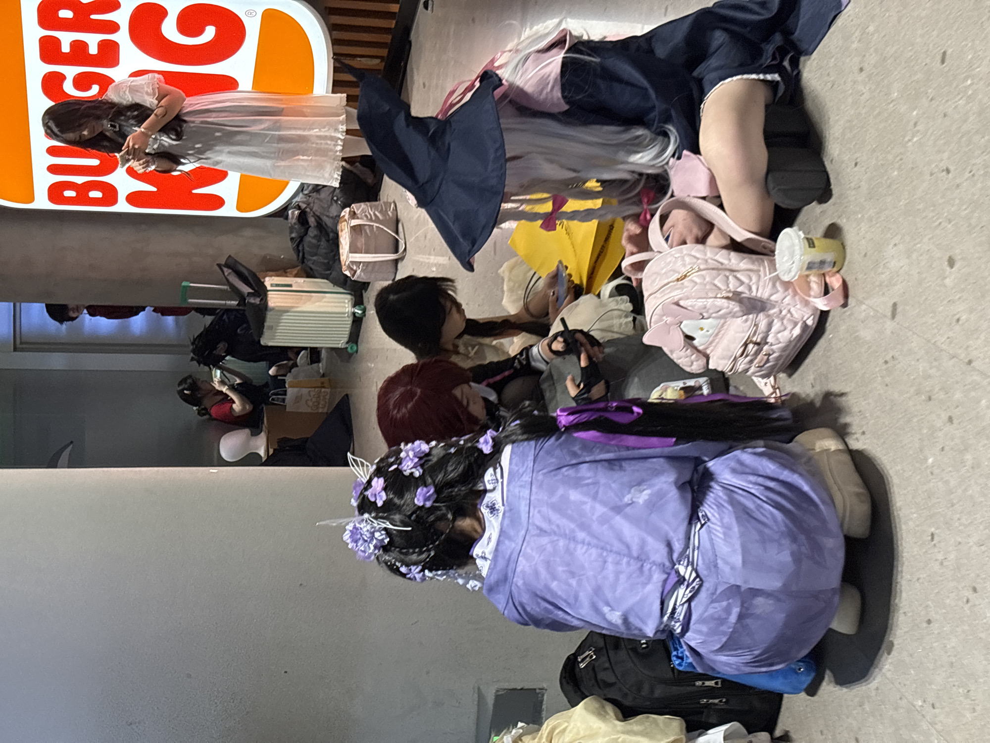 The scene captures a gathering of individuals dressed in cosplay, possibly at a convention or event. The location appears to be a sidewalk in an urban setting. Costumes suggest a theme, possibly anime or gaming-related. Several people are seated on the ground surrounded by luggage, including suitcases and bags. A Burger King sign is visible in the background. The composition suggests a break or resting period during an event.