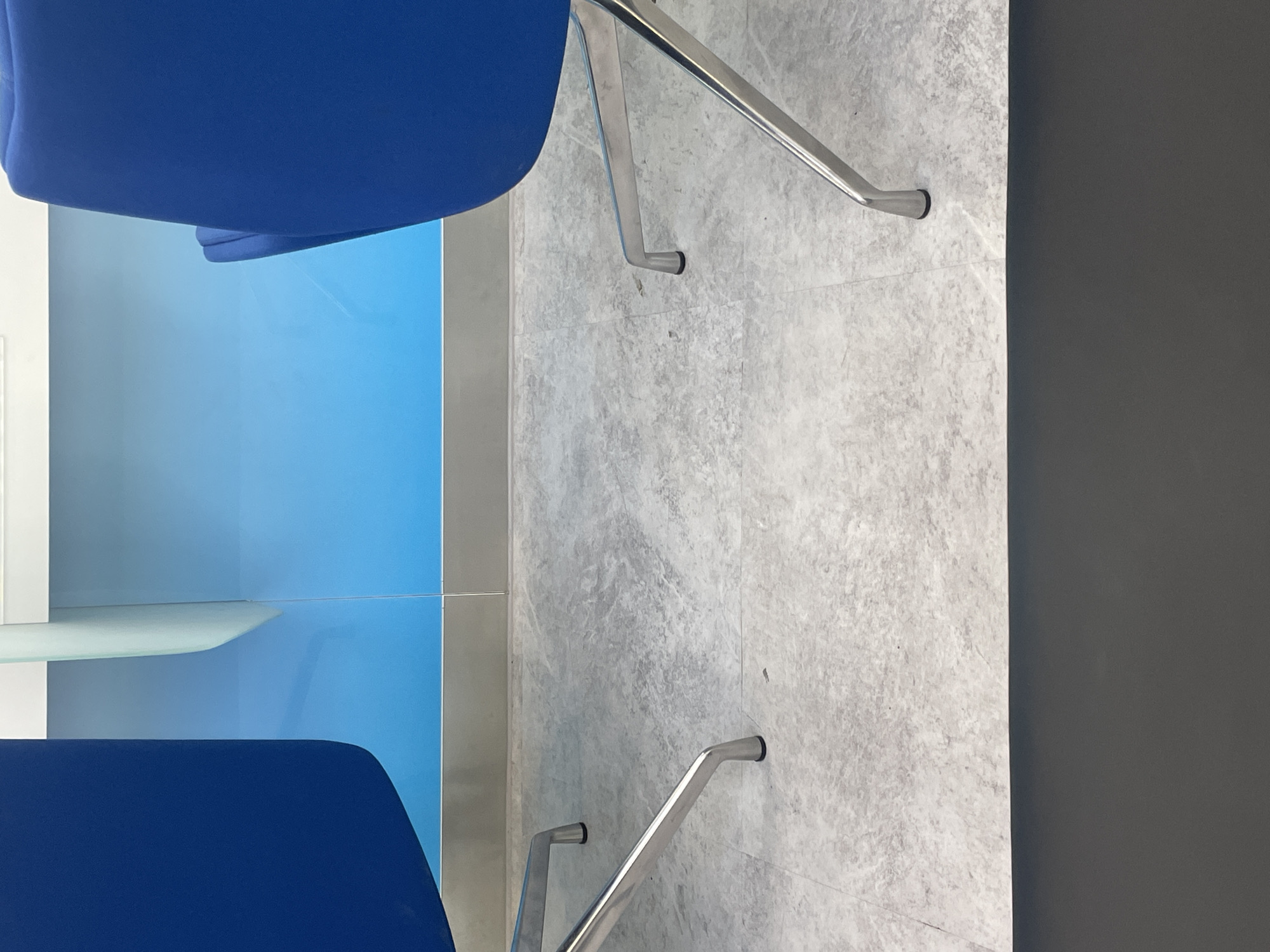 The image depicts the lower half of two blue chairs with metallic legs, positioned on a grey marble-patterned floor. Behind the chairs, a blue wall with a subtle gradient extends upwards, meeting a white horizontal surface. The scene is devoid of any human presence or activity. The lighting suggests an indoor environment, possibly an office or waiting area. The specific location in Ulaanbaatar, Mongolia, cannot be determined from the visual information provided.