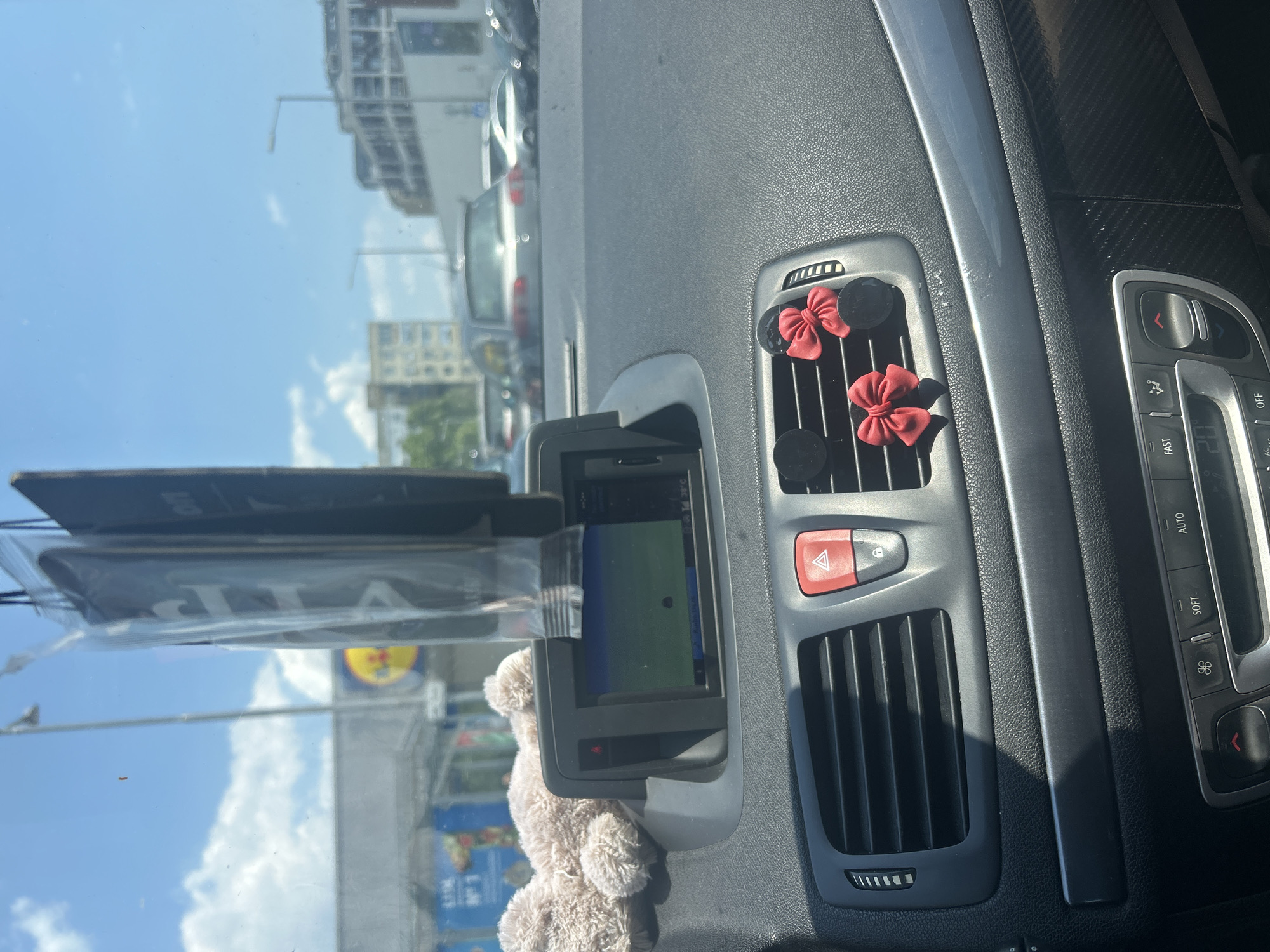 The image shows the interior of a car, looking out through the windshield on a sunny day. The car dashboard is visible, with a navigation screen showing a map, air vents, and various buttons. There are two small red bows attached to the air vents, and a fluffy toy is resting on the dashboard. Outside the car, buildings, streetlights, and other vehicles can be seen, suggesting the car is in an urban area. A sign for the supermarket Lidl is visible in the background, suggesting the location may be near a Lidl store. While the image does not definitively place the car in Plovdiv, Bulgaria, the presence of a Lidl supermarket is consistent with many urban environments in Bulgaria. The scene captures a moment of everyday life, possibly during a car journey or while parked in a city.
