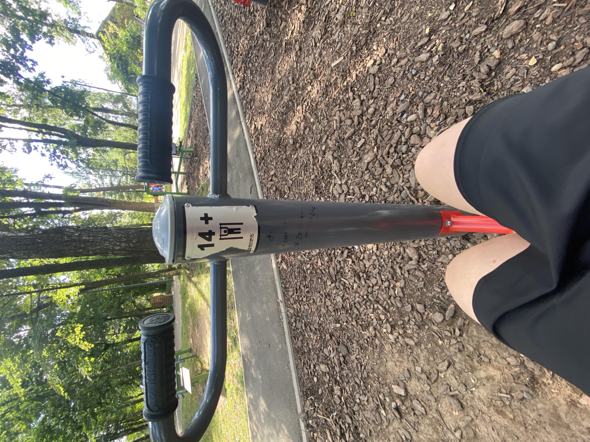 The image shows an outdoor fitness machine in a park on a sunny day. The machine has a central grey pole with a "14+" sign and a diagram of a person doing pull-ups. The handles of the machine are black and textured. In the foreground, a person's legs in black shorts are visible, sitting on the machine. The ground is covered in wood chips and small stones. In the background, there are trees with green foliage, a paved path, and other fitness equipment. The location is Votkinsk, Russia, as indicated by the text on the machine which includes "VOTKINSK". There are some graffiti markings on the grey pole below the "14+" sign.