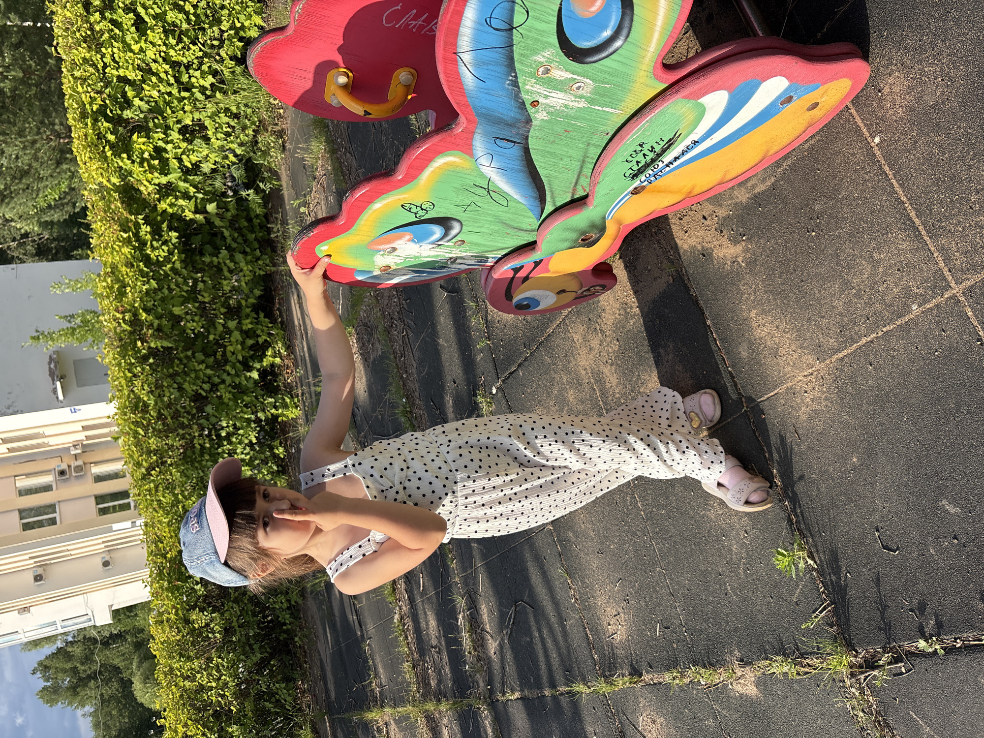 A young girl in a white polka-dot jumpsuit and a blue and pink baseball cap stands on a playground. She is positioned to the left of a colorful, butterfly-shaped playground ride, which is primarily red and green with yellow and blue accents. The girl is looking towards the viewer, with her right index finger raised to her nose, and her left hand resting on the butterfly ride. The playground surface is made of dark rubber tiles, with grass growing through the cracks between them. In the background, there is a neatly trimmed green hedge, and a light-colored, multi-story building with windows and air conditioning units. The lighting suggests it is a sunny day. The image is captured in a city park or playground in Chaikovsky, Russia.