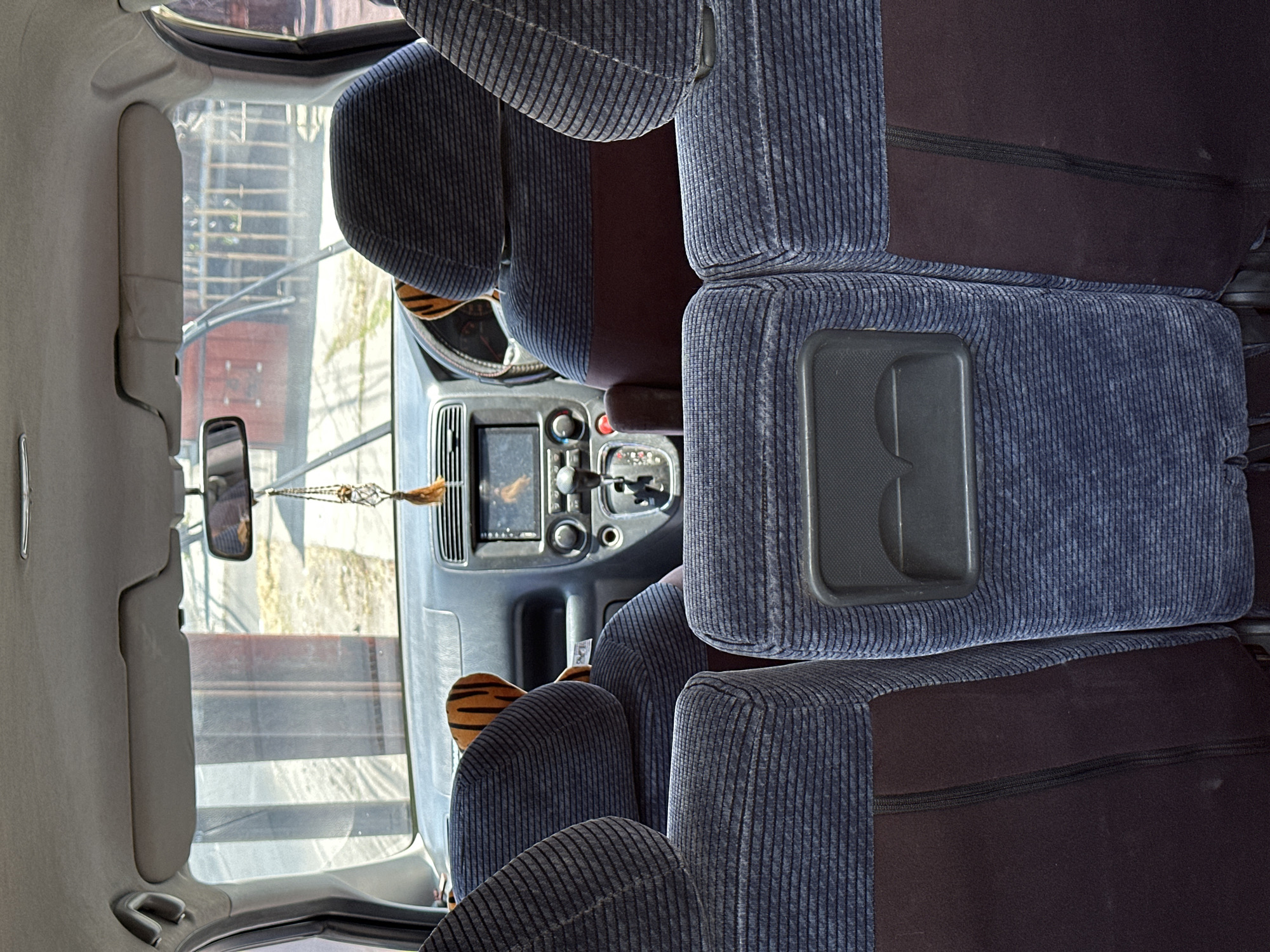The image, taken from the rear passenger seats of a car in Bulakrejo, Indonesia, provides a view of the car's interior and the outside environment through the windshield. The car's interior features blue upholstered seats with a dark red accent on the seat backs. A stuffed tiger toy is visible on the dashboard, and a decorative beaded ornament hangs from the rearview mirror. The dashboard includes a car stereo and gear shift. Outside the car, a building with a red facade and a metal fence is visible, along with some greenery and a paved area. The windshield wipers are down, suggesting the car is stationary.