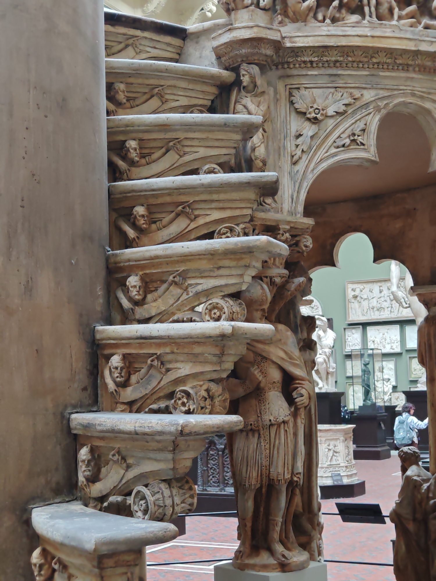 The image shows a section of the Victoria and Albert Museum in London, United Kingdom. The foreground features a large, intricately carved stone structure. On the left, a vertical element supports horizontal steps, each adorned with human figures and decorative elements. To the right, a partial archway is visible, with sculpted figures and floral motifs. Through the archway, the museum's interior is visible, displaying various sculptures and reliefs. A person wearing a light blue jacket with a backpack is seen near the statues in the distance. The flooring is red and white tiled.