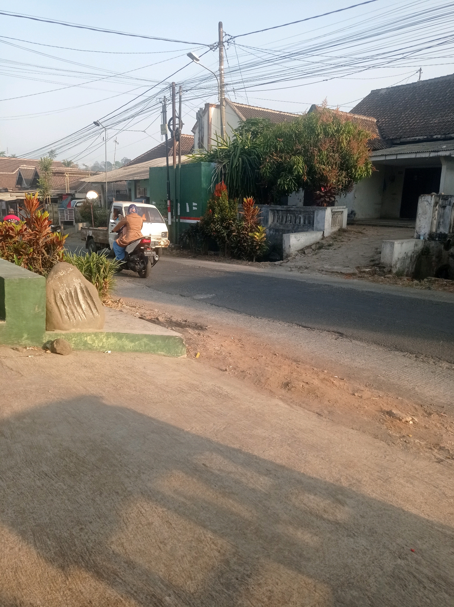 In Purwosekar, Indonesia, a person rides a motorcycle down a paved road lined with residential buildings and vegetation. A small white truck is parked nearby. Overhead, a dense network of utility wires crisscrosses the sky, supported by utility poles. In the foreground, a concrete sidewalk and a large, sculpted rock are visible. The scene is bathed in the warm light of what appears to be early morning or late afternoon.