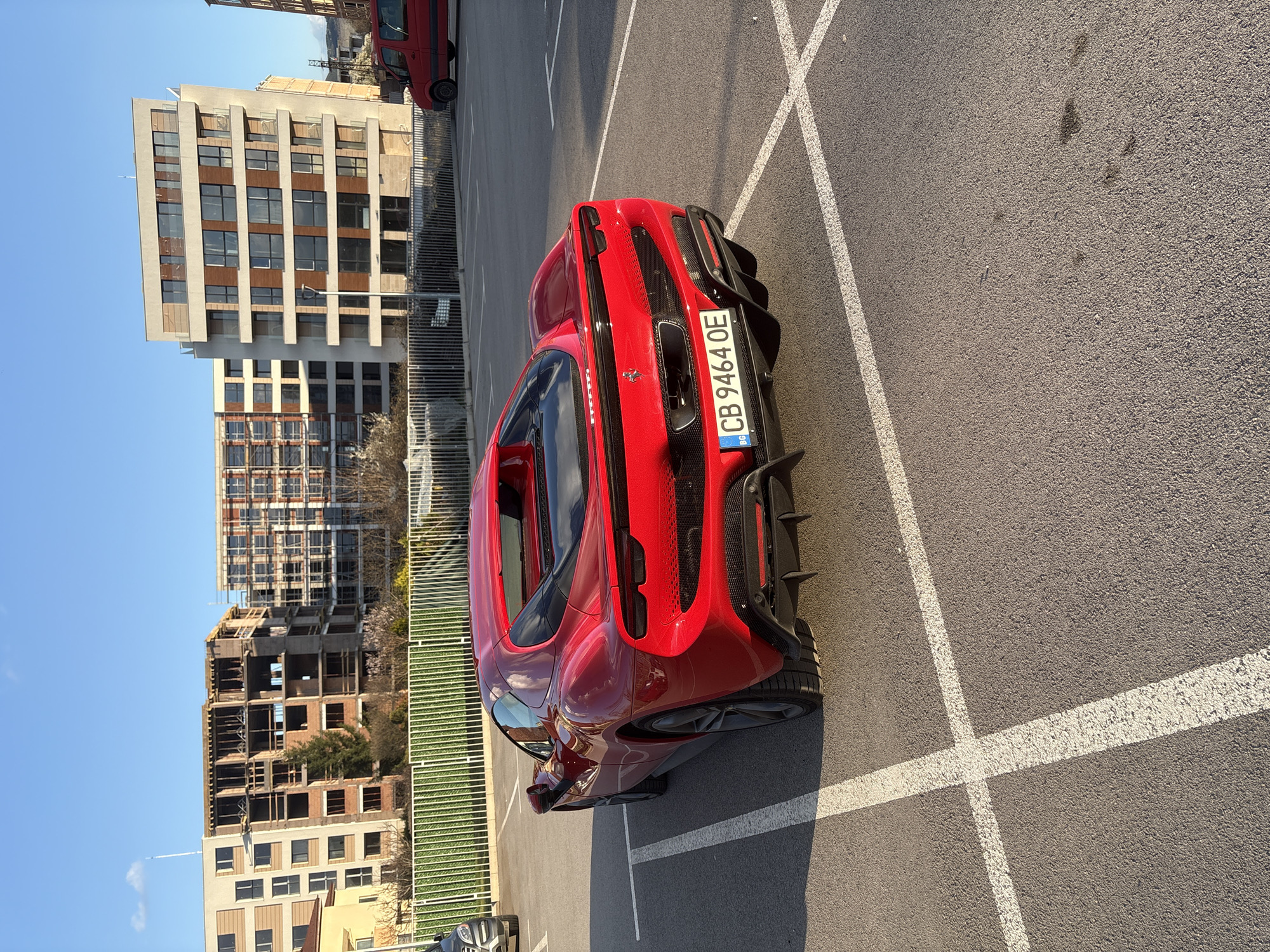 The image shows a red Ferrari parked in a parking lot with white parking space lines. In the background, there are several buildings under construction and completed buildings. The license plate on the car reads "CB 9464 OE," indicating that the car is registered in Bulgaria. Given the type of license plate and the urban environment with visible construction, the location is likely in a major city, which aligns with the context clue that the city is Sofia, Bulgaria. There are no visible people or interactions in the scene. The lighting suggests it is daytime with clear weather. A red van is barely visible in the background near the top edge of the photo.