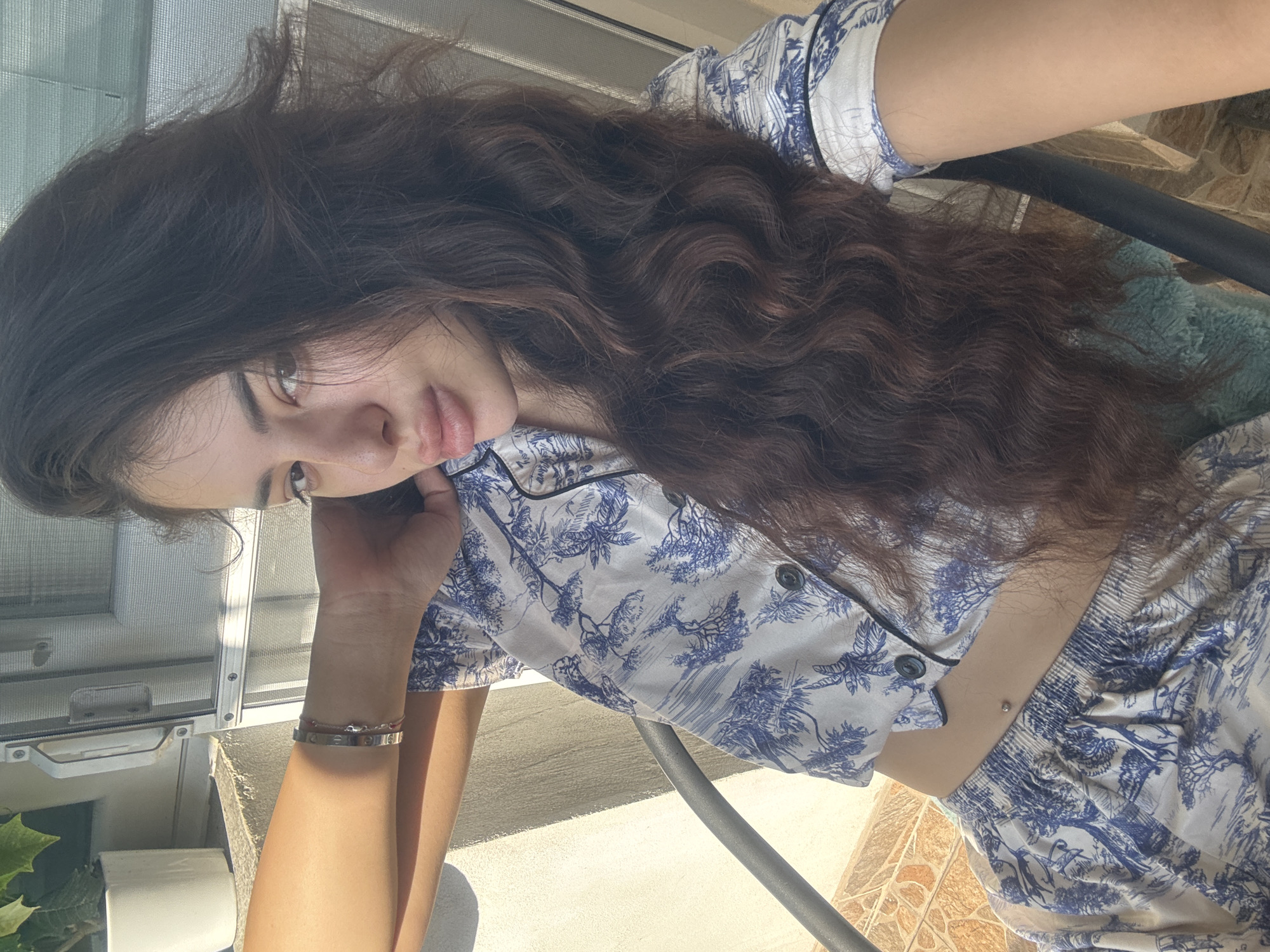 A young woman with long, dark, wavy hair is captured in a close-up, outdoor portrait. She is seated, leaning back with her left arm resting on her head and her right arm bent, with her hand near her face. She is wearing a pajama set with a blue and white toile pattern featuring trees and foliage. A silver bracelet is visible on her left wrist. Her skin is tanned, and she has noticeable full lips and dark eyes. A navel piercing is visible on her abdomen.
The background suggests an outdoor setting, likely a balcony or patio. A textured wall, possibly concrete, is visible to the left, with sunlight casting shadows. A white planter with green foliage is on a ledge above. To the right, a screen door with a metal frame and handle is partially visible. The flooring appears to be tiled. The overall lighting indicates a sunny day. The scene does not contain any specific landmarks or clues that would allow for a precise identification of the location within Plovdiv, Bulgaria.
