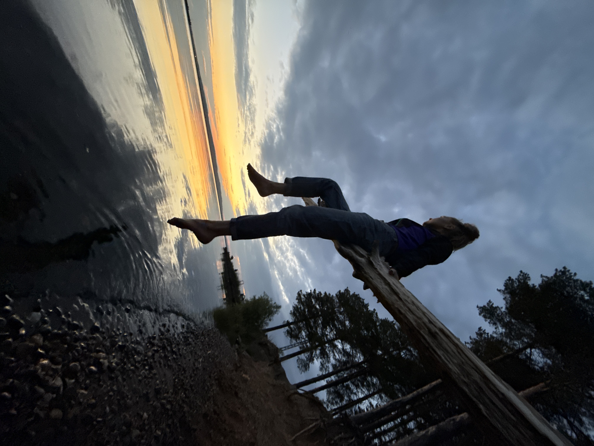 A young woman with her hair in a bun sits on a fallen tree trunk extending over a body of water at dusk. The sky is cloudy with a band of orange and yellow sunset colors visible on the horizon. The water reflects the sky and the surrounding trees. The trunk is positioned diagonally in the frame, with the woman seated on it facing away from the camera, looking towards the sunset. Her legs are dangling over the water. The shoreline to the right of the frame is covered in dark pebbles and bordered by a slope with trees. The location is the Kama River in the city of Chaikovsky, Russia.