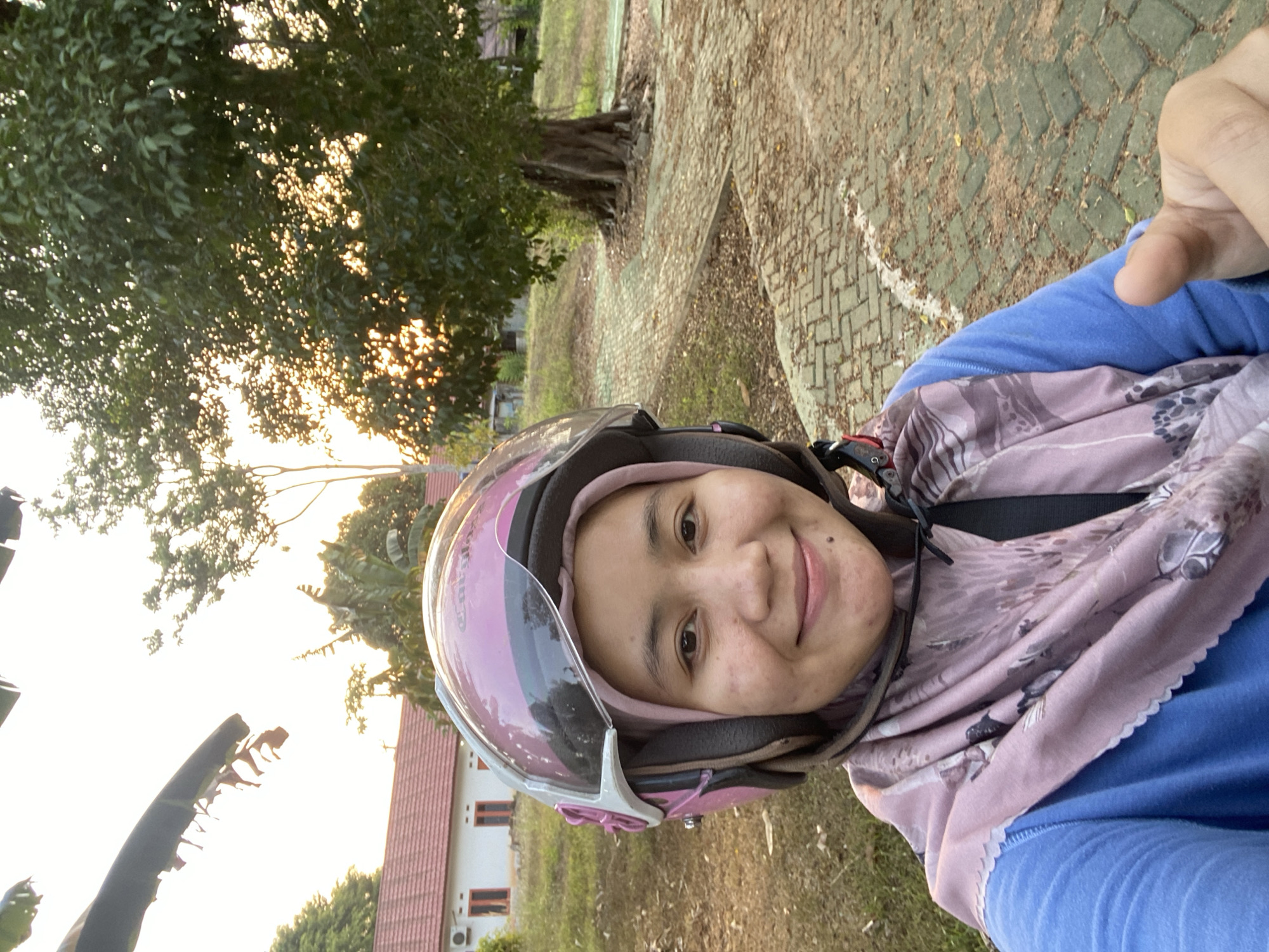 A young woman wearing a pink helmet and a blue shirt with a patterned scarf is taking a selfie outdoors. The background shows a building with a red roof, a large tree, and a paved walkway. The setting appears to be a residential area or a park in Banjarbaru, Indonesia, given the vegetation and architecture. The lighting suggests it is late afternoon or early evening, with the sun setting behind the trees. The woman is smiling and giving a thumbs-up gesture.