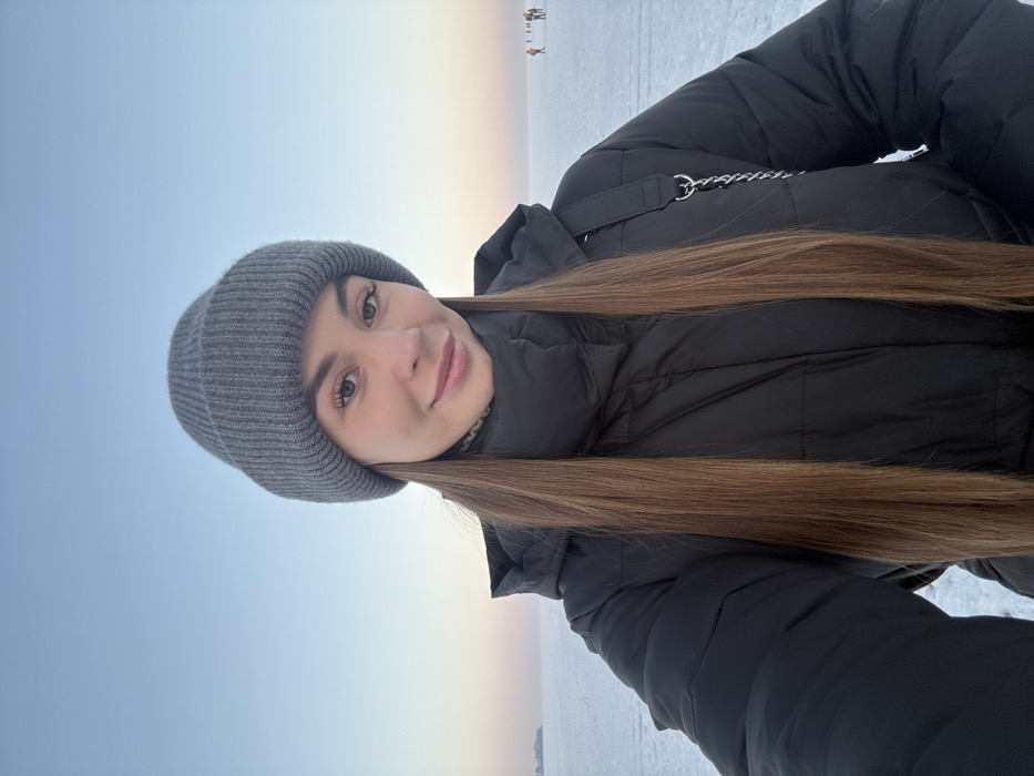 A person in a gray beanie and a black puffer jacket takes a selfie outdoors. The sky is a pale blue with a hint of pink near the horizon. A flat, snowy expanse stretches out behind the person, with a few small figures visible in the distance. The person has long, straight brown hair that falls across their chest. They have light skin, brown eyes, and a slight smile. A silver chain is visible on the jacket.