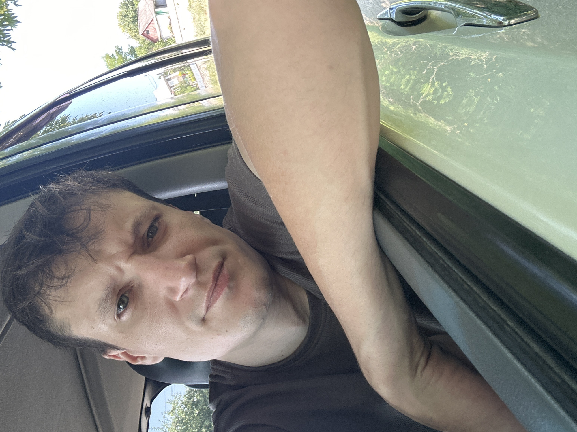 The image is a selfie taken from inside a vehicle, looking towards the driver's side. A person with light skin, dark hair, and green eyes is visible. They are wearing a dark gray t-shirt and have their left arm resting on the open car door, with their elbow and forearm clearly visible. The background outside the car shows greenery, trees, and a few buildings, suggesting a rural or suburban setting. The car appears to be light green in color, with a chrome door handle visible on the right side of the frame. The lighting suggests it is daytime. The person's facial expression is a slight grimace. No specific landmarks identifying the city of Makiv, Ukraine are present in the image.