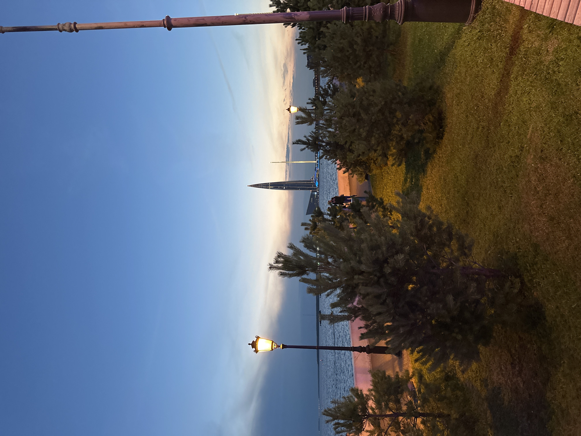 This is a photograph taken at dusk, likely from a waterfront park or embankment in Saint Petersburg, Russia. The sky is a gradient of deep blue at the top transitioning to lighter hues of orange and pink near the horizon, with some scattered clouds.
In the foreground, there is a grassy area with several small evergreen trees. A lamppost with a glowing orange light stands to the left of the frame, and another ornate lamppost is partially visible on the far right.
In the middle ground, a body of water, likely the Gulf of Finland or a section of the Neva River, stretches across the scene. On the far side of the water, the distinctive Lakhta Center, a prominent skyscraper in Saint Petersburg, is visible, illuminated with blue lights. A lighter-colored, slender tower is situated next to it. A few people can be seen walking along the embankment on the other side of the water.
The lighting suggests it is twilight, with artificial lights from the lampposts and the skyscraper illuminating the scene. The overall atmosphere is serene and urban.