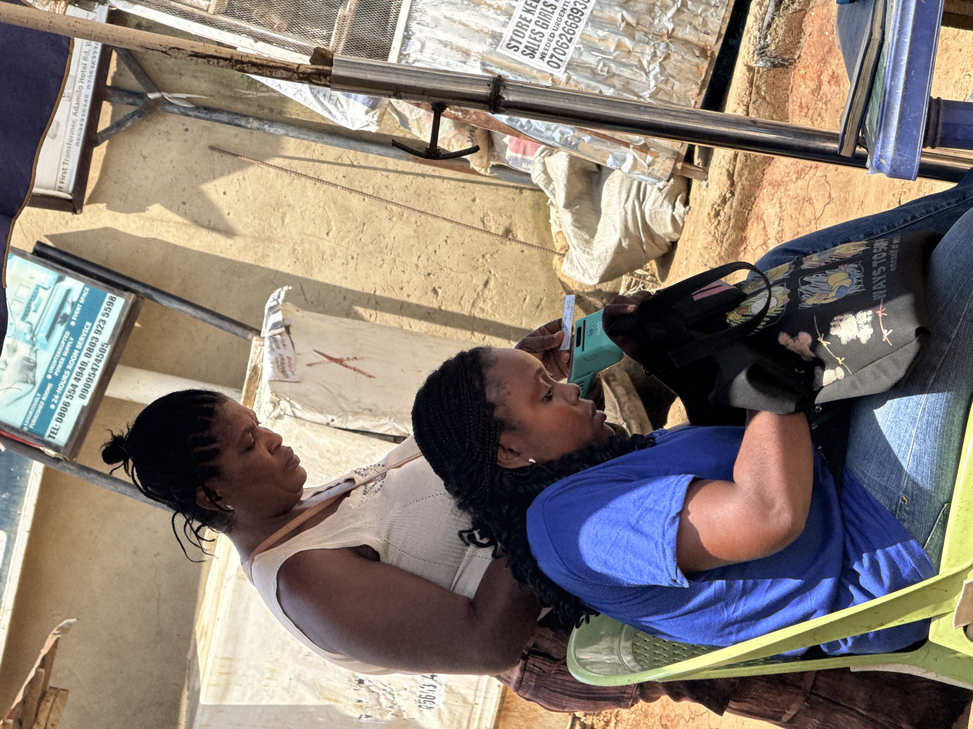 In Abuja, Nigeria, a woman with braided hair and wearing a white sleeveless top is standing behind another woman who is seated on a green plastic chair. The seated woman, also with braided hair, is wearing a blue t-shirt and denim jeans. She is holding a small green electronic device and a black bag with cartoon characters and text, which appears to have the words "WAYS TO SU" visible. The background shows a sandy, sun-drenched environment with corrugated metal structures and sacks of goods. A partially visible sign with text and numbers is also present. The lighting suggests it is daytime.