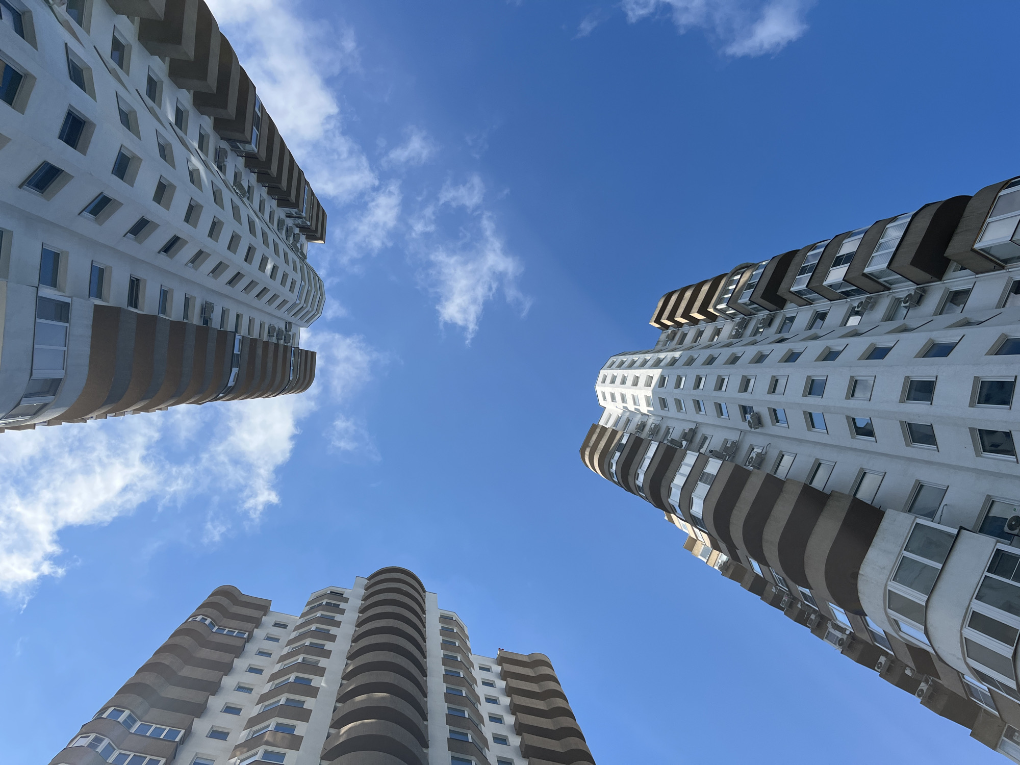 This is a low-angle, wide-shot photograph of three modern apartment buildings against a clear blue sky with scattered white clouds. The buildings are tall and appear to be residential, with multiple floors and balconies. One building on the left and another on the right are taller and more prominent, reaching towards the top of the frame. A third, shorter building is visible at the bottom center. All buildings have a similar architectural style, featuring white facades, dark-framed windows, and curved, projecting balconies with a warm, neutral color. The balconies are stacked in a way that creates a rhythmic pattern along the building's exterior. Some of the buildings have visible air conditioning units mounted on their walls. The perspective from below emphasizes the height and scale of the structures, making them appear to lean into the sky. The lighting suggests it is daytime, likely midday, given the bright sky and clear visibility. There are no people clearly visible in the image, and no text is present. The setting appears to be an urban residential area, possibly in Sochi, Russia, based on the provided location context, characterized by contemporary architecture and open sky. The overall mood is one of architectural dominance against a serene sky.