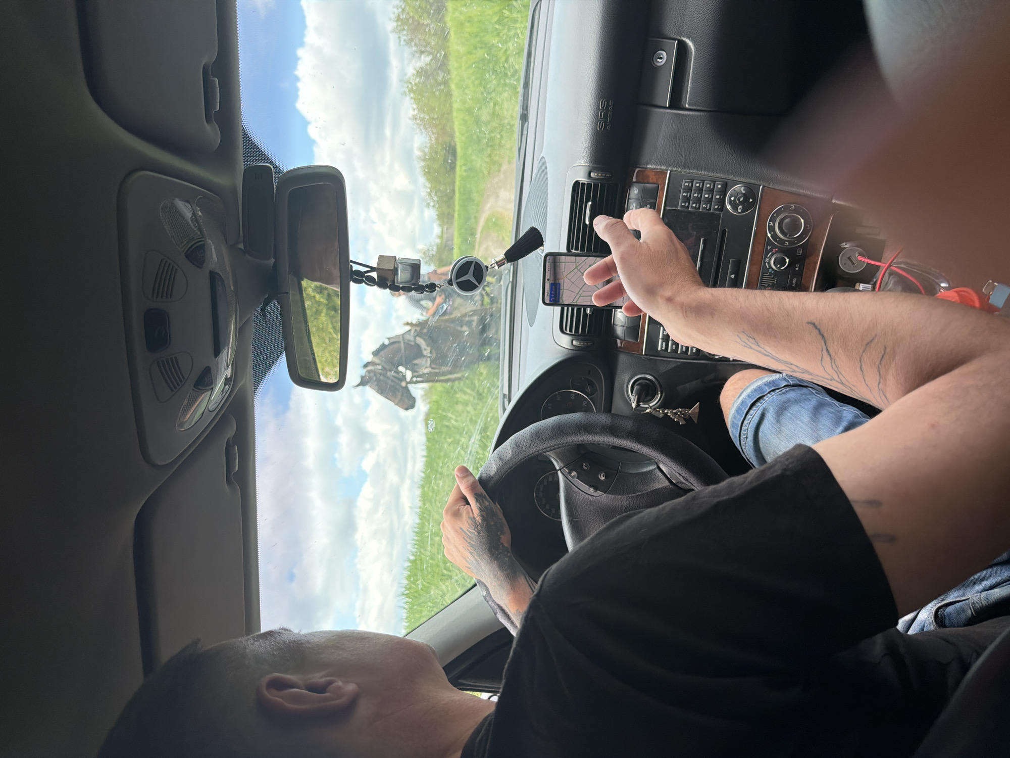 A person is driving a Mercedes-Benz. The driver is using a mobile phone for navigation. The location appears to be in a rural area in Leningradskaya, Russia. A crash scene is visible in the car's rear-view mirror. The driver is wearing a black t-shirt and blue denim shorts. The driver has tattoos on their arms.