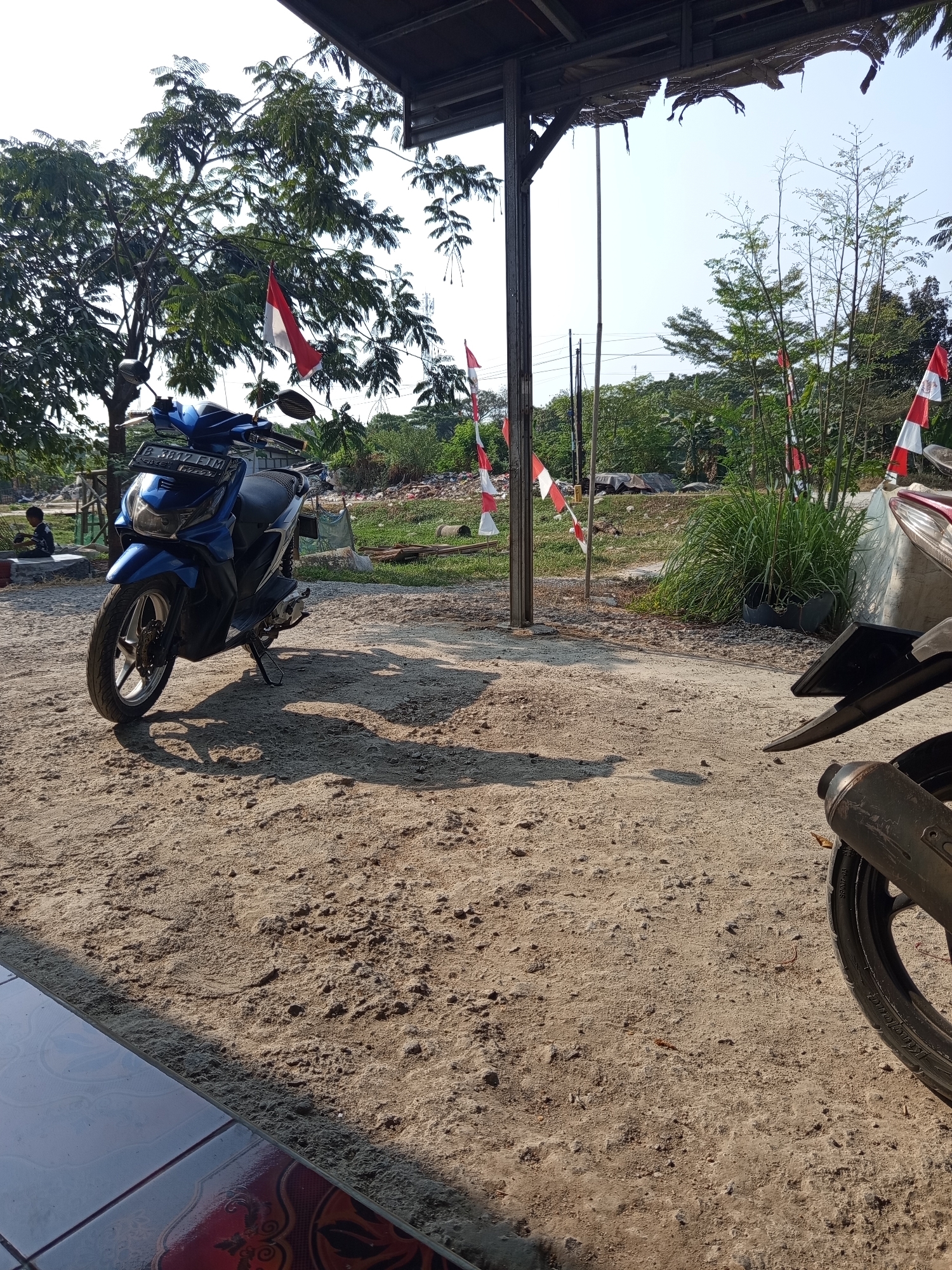 A blue scooter is parked on a sandy area under a covered structure. Red and white Indonesian flags are visible in the background. The scooter has a license plate that reads "B 3887 FJM". A person is sitting on a low wall in the distance. The scene appears to be in an outdoor, semi-rural setting.