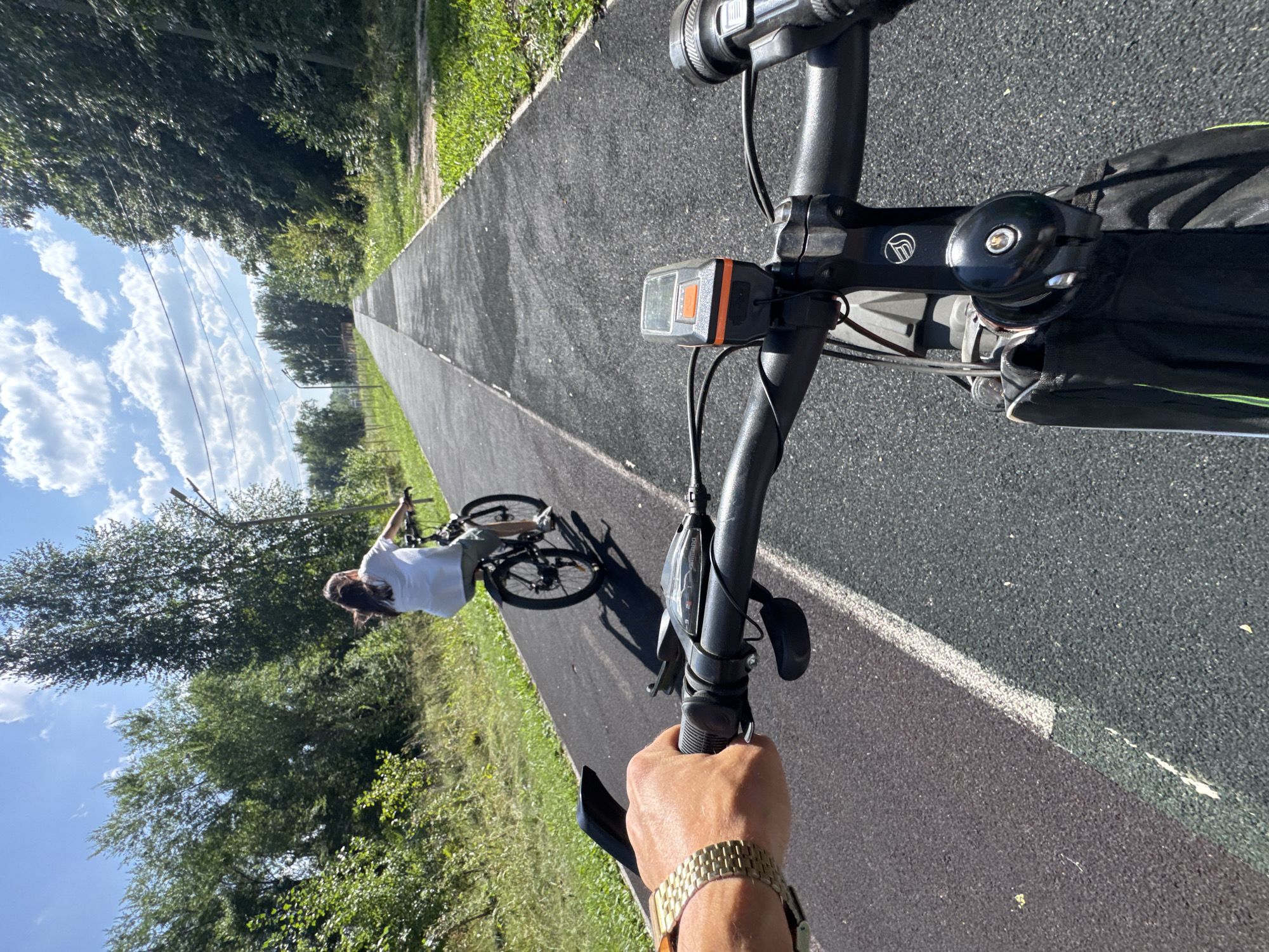 In Oskemen, Kazakhstan, on a sunny day, two people are cycling on a paved path lined with trees and grass. The perspective is from the handlebars of the leading bicycle, showing the rider's left hand with a gold watch. Ahead, another cyclist, a person with long dark hair wearing a white t-shirt and shorts, is riding a black bicycle. The path appears to be a dedicated bike lane, with a white line marking its edge. The sky is blue with scattered clouds, and the overall environment suggests a park or recreational area within the city.