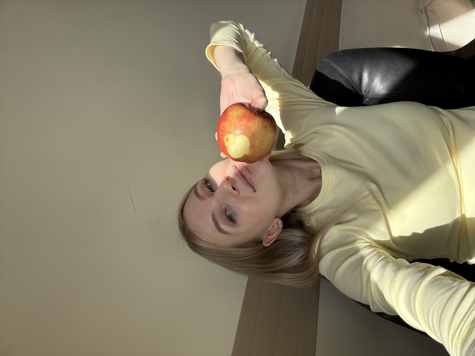 A fair-skinned young woman lies on a black, leather-like surface, possibly a couch or chair. She is wearing a long-sleeved, light yellow shirt. She holds a partially eaten red apple above her face. Her hair is blonde, and she has blue eyes. The background is a light-colored wall illuminated by sunlight. Based on the available information, it's not possible to determine if this image was taken in Izhevsk, Russia.