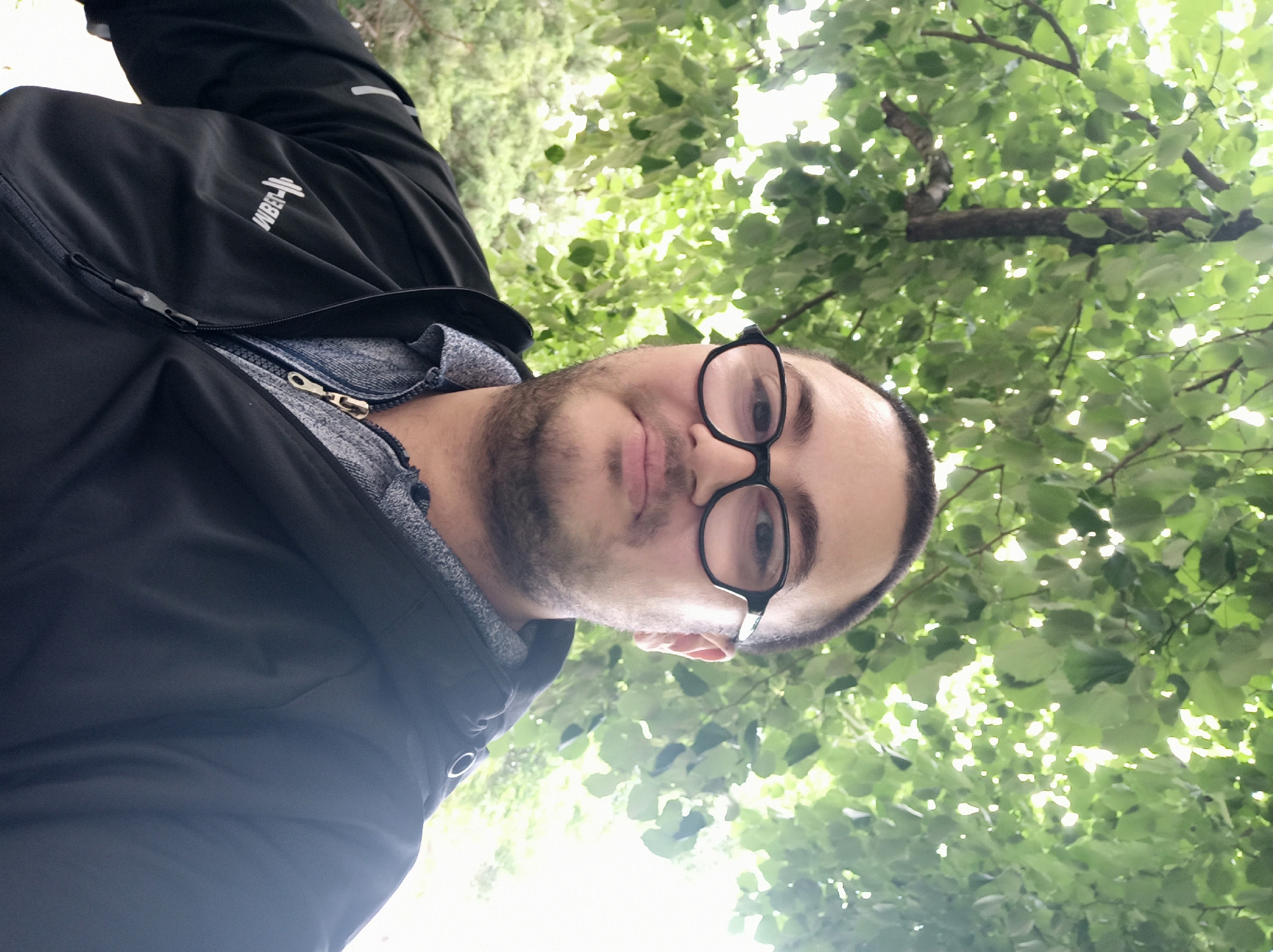 A person wearing glasses and a black jacket is pictured in front of green foliage, possibly trees, in an outdoor setting. The location is Unknown, Unknown.