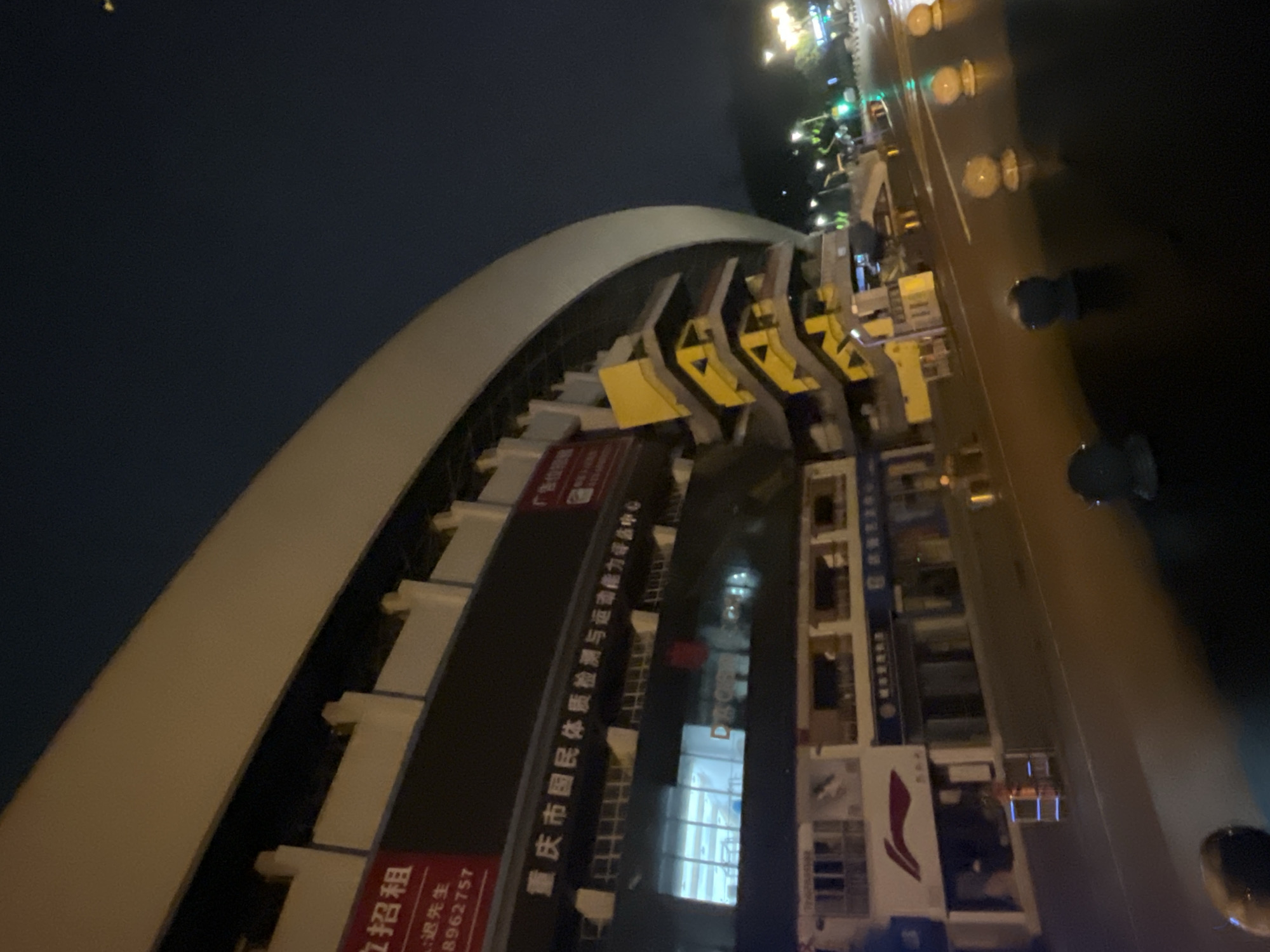 The image captures an exterior night view of a large, multi-story building featuring a distinctive curved, light-colored roof structure. The scene is illuminated by artificial lights.
The building, identified as the Chongqing National Physical Fitness Testing and Sports Ability Assessment Center ("重庆市国民体质检测与运动能力评估中心" visible in white Chinese characters on a dark facade), is located in the Jiulongpo District of Chongqing, China. Red signage on the building also displays "正在招租" (For Rent) and "广告位" (Advertisement space), along with a partial phone number "8962757". Below the main facility, street-level businesses are visible, including one with a red "Li-Ning" (李宁) sportswear logo. A prominent yellow, multi-tiered external staircase system is affixed to the right side of the building.
In the foreground, a dark asphalt road surface reflects light, suggesting it is wet. Several light-colored, spherical road bollards are evenly spaced along the road, alongside a few darker, rounded bollards. Distant green traffic lights and other street lights are visible, indicating an active urban environment. No individuals are discernible in the frame. The sky above is dark, indicating night conditions.