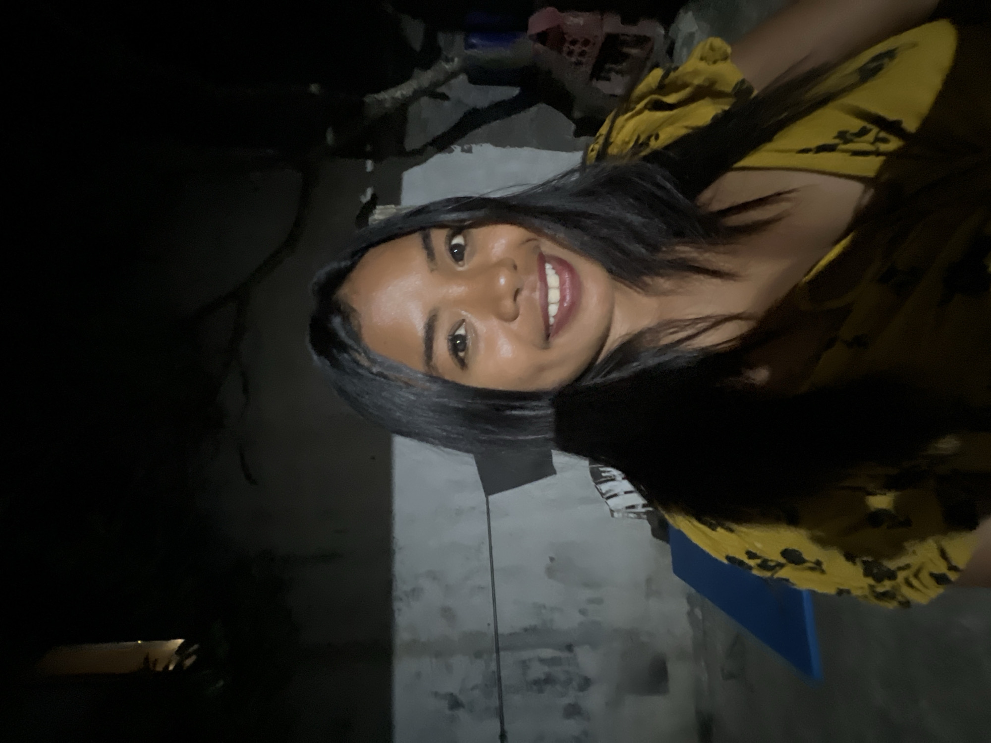 The image is a close-up, selfie-style photograph of a young woman with dark hair and brown eyes. She is smiling and wearing a yellow dress with a black floral pattern. The background is dark and appears to be an outdoor setting at night. A textured, light-colored wall is visible behind her, with some dark shapes that could be foliage or structures. To the left of the woman, a blue object, possibly a mat or cushion, is partially visible on the ground. In the background, there are some indistinct structures and branches of trees. The lighting suggests an artificial light source is illuminating the woman. The scene is likely a residential area in Denpasar, Indonesia, given the context.