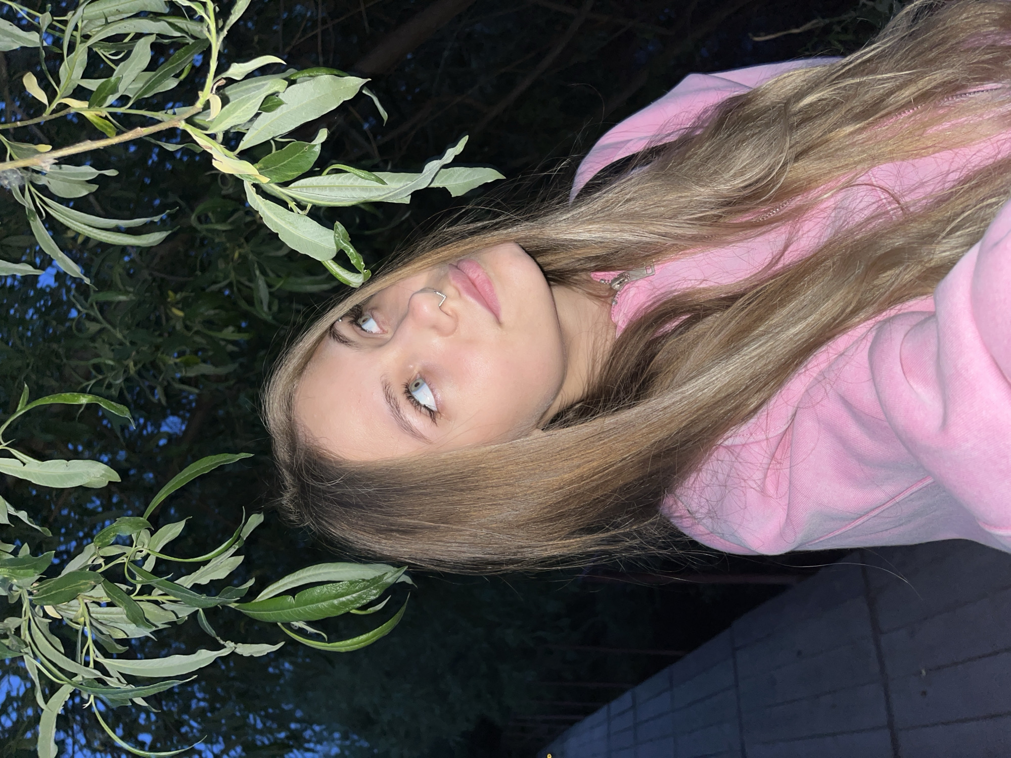 A young woman with long, blonde hair is facing the camera. She is wearing a pink hoodie with the zipper partially visible. She has fair skin, light-colored eyes, and a nose piercing. A leafy branch is in the upper left of the image. The background is dark, suggesting it is night time. The setting appears to be outdoors near a wall or building. The image location is assumed, as Фоки, Russia, is not mentioned in the image.