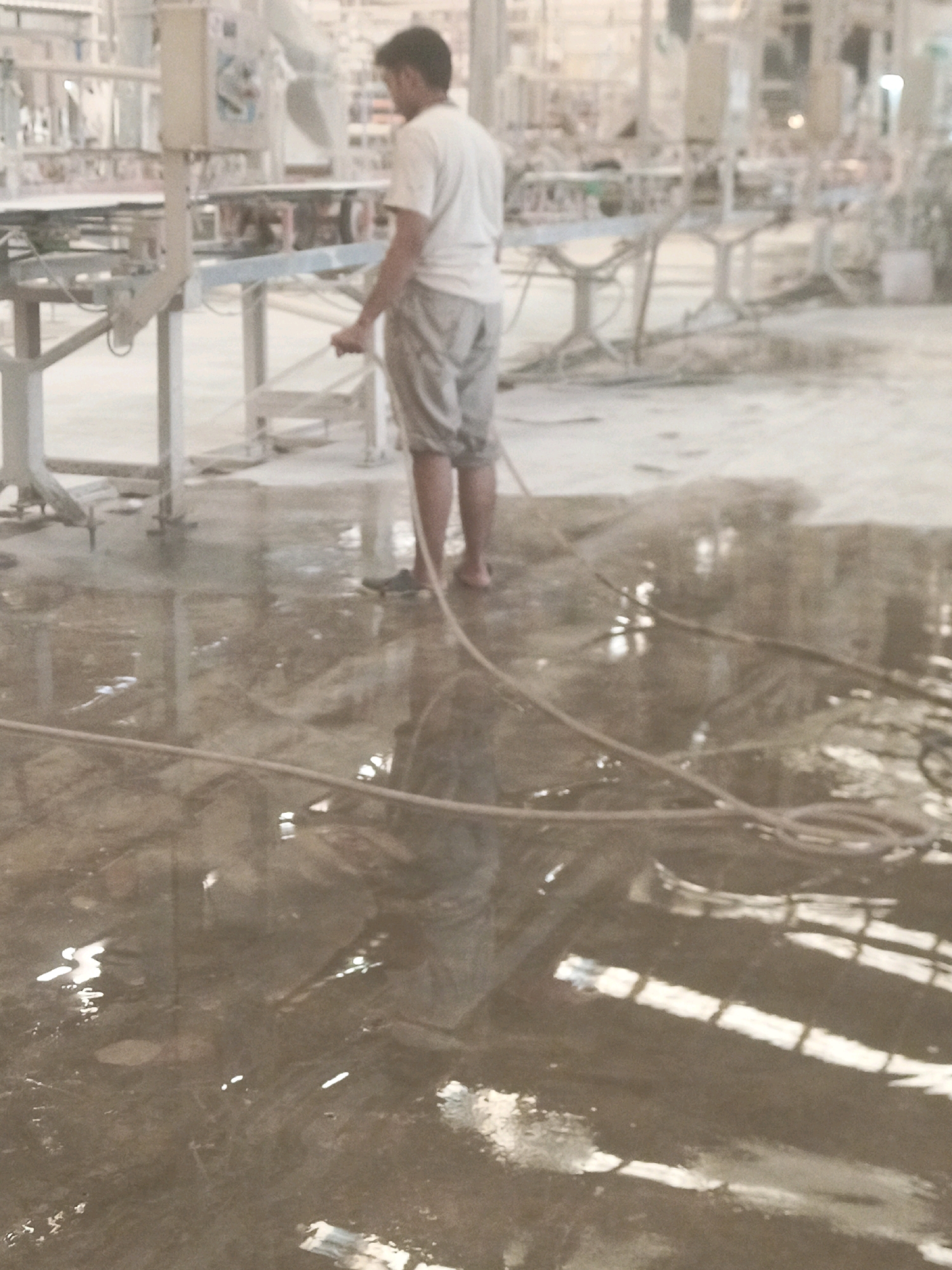 An Indonesian man, wearing a white t-shirt and grey shorts, stands in a flooded industrial area in Lebaniwaras, Indonesia. He is holding a hose connected to a spraying apparatus, suggesting he is engaged in cleaning or a similar task. The floor is covered in water, reflecting the overhead fluorescent lights and the surrounding industrial machinery. The environment appears to be a factory or workshop, with various metal structures and equipment visible in the background. The water on the floor creates ripples and distortions, obscuring some of the details of the machinery and the ground.