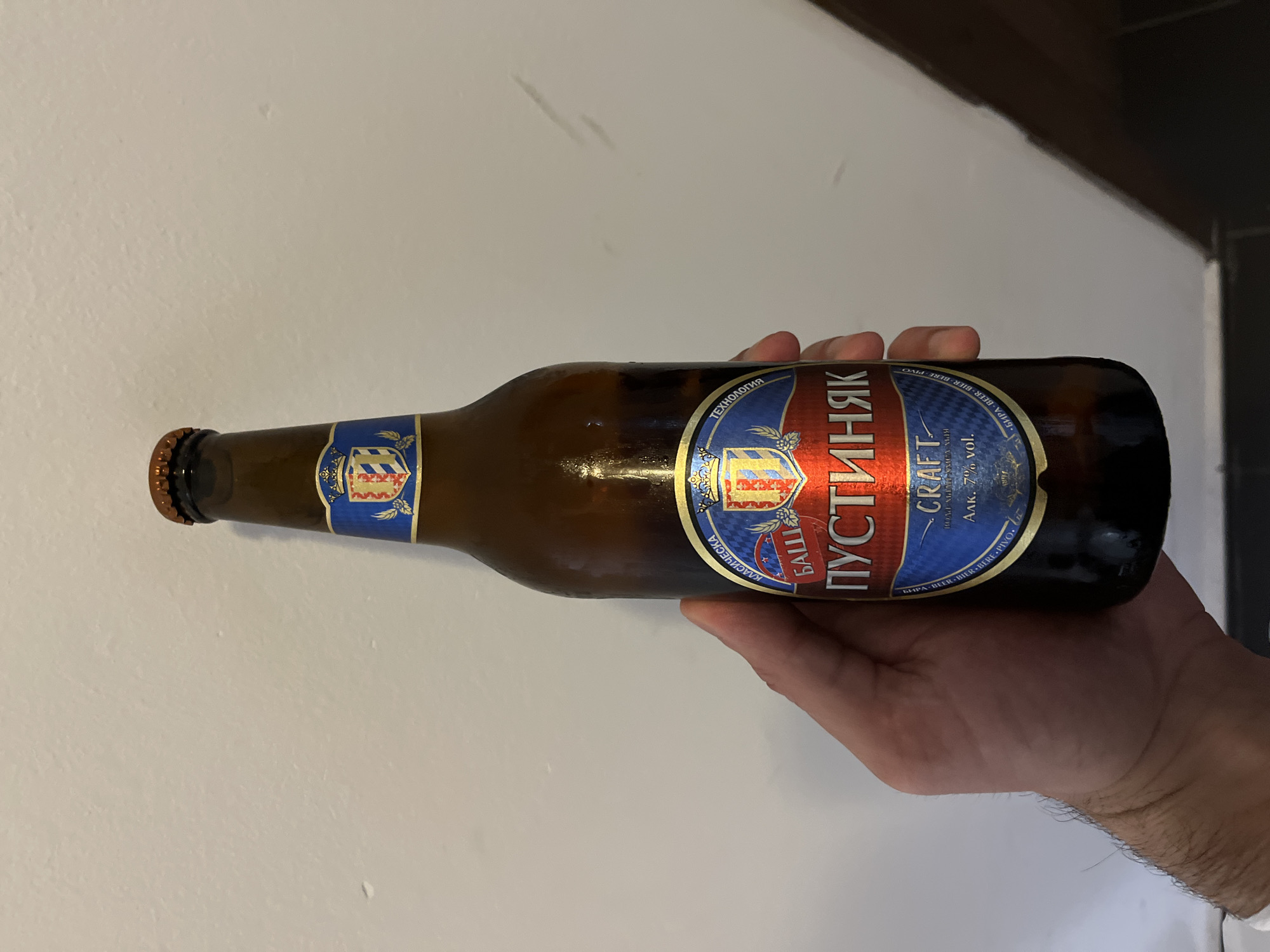 A hand holds a bottle of "Bashi Pustinyak" craft beer against a white wall. The bottle has a blue and red label with gold lettering and a crest. The background is a plain white wall, and the lighting suggests an indoor environment. There are no visible city landmarks or indicators of Sofia, Bulgaria. The image focuses solely on the beer bottle and the hand holding it.