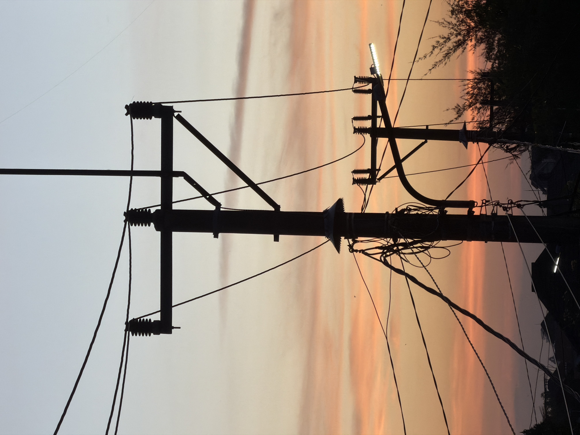 The image shows two utility poles silhouetted against a sunset sky. The sky has hues of gray, white, orange, and pink. The left pole is simpler in design, with three horizontal supports holding insulators and wires. The right pole is more complex, with numerous wires and insulators, and a small light fixture attached at the top. Trees or shrubs are silhouetted behind the right pole. The location is North Kuta, Indonesia.