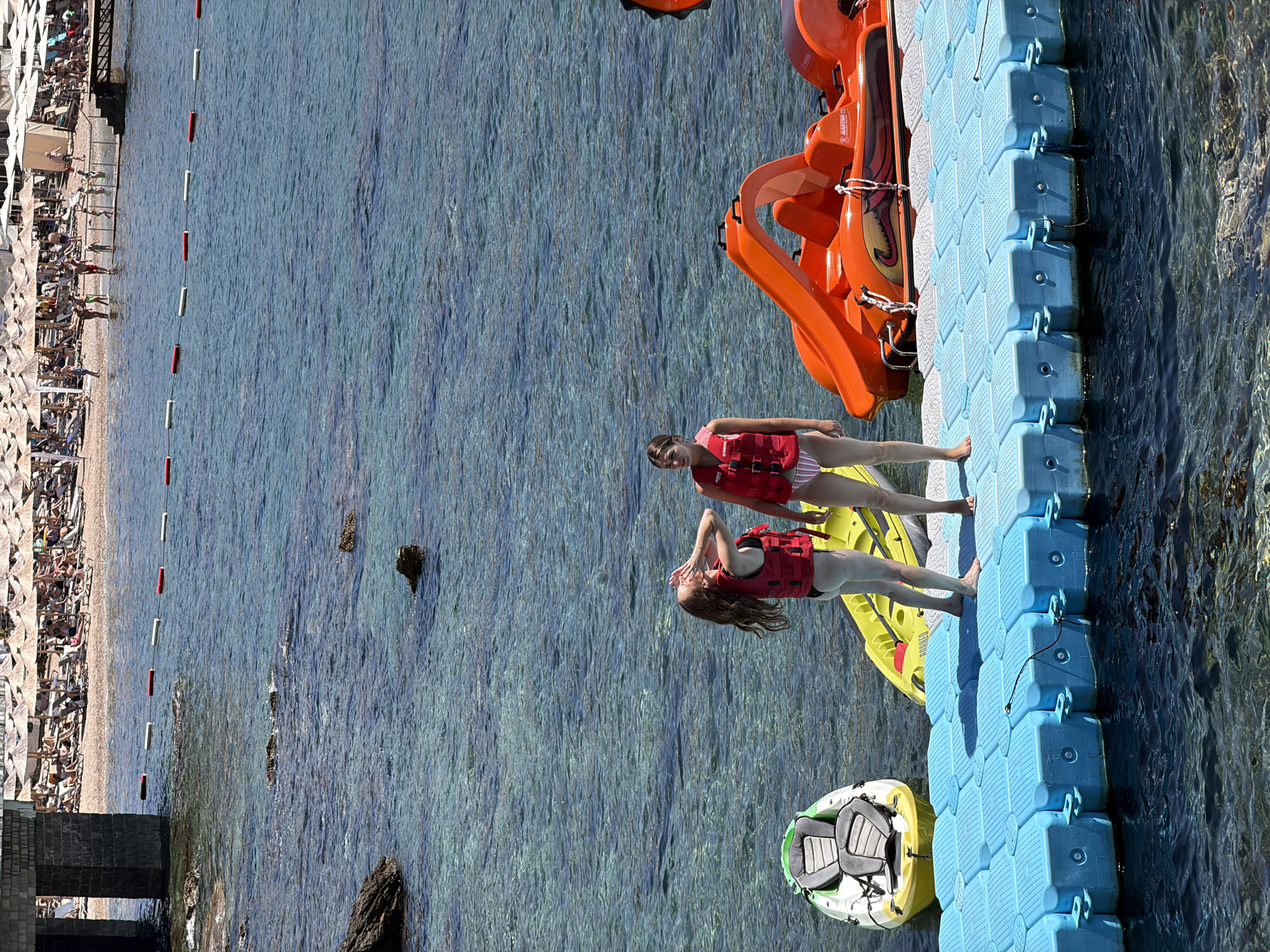 Two young women are standing on a blue floating dock in the turquoise waters of the Adriatic Sea, just off the coast of Budva, Montenegro. They are both wearing red life jackets and swimwear. One woman, on the left, has long, reddish-brown hair and is holding her hands up to her face. The other woman, on the right, has dark hair pulled back and is looking to her left.
Beside them on the dock are a white and green kayak and a bright yellow kayak. Further to the right is a large, orange pedal boat with a slide. In the background, a crowded beach is visible, with rows of sun loungers and thatched umbrellas. The water is clear, allowing a view of the rocky seabed. A line of buoys separates the swimming area from the open water. The scene suggests a sunny day at a popular tourist destination.