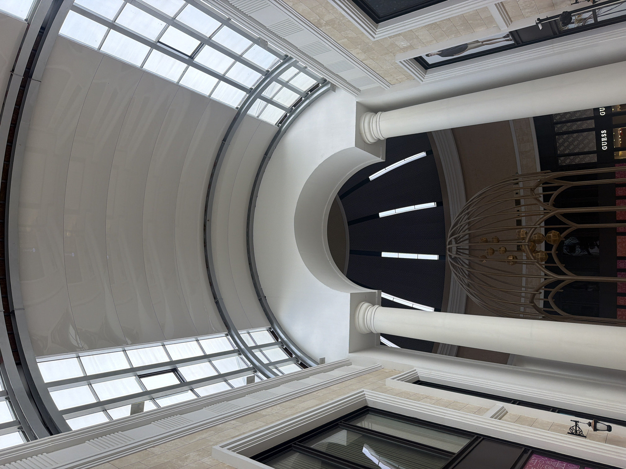 An interior shot captures a grand atrium with a domed ceiling and a skylight made of glass panels. The atrium features large white columns and decorative architectural elements on the walls. Below the dome, a decorative metal structure with hanging spheres is visible, and a store with the "GUESS" logo is present. The scene is illuminated by natural light from the skylight and artificial lighting from within the atrium. No people are visible in the image. This image was taken in Serik, Turkey, specifically at the Land of Legends shopping area, as identified by the "GUESS" store and the overall architectural style commonly found in that location.
