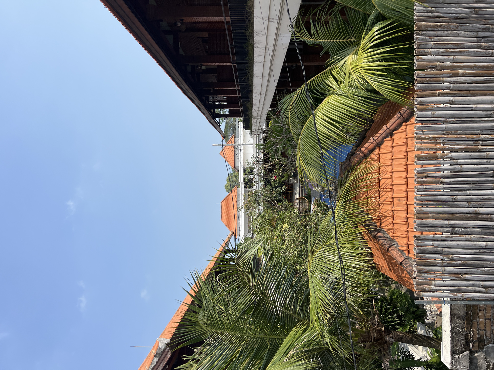 The image shows a view of a tropical setting, possibly a resort or residential area, in North Kuta, Indonesia. In the foreground, a fence made of vertical bamboo poles is visible. Behind the fence, lush green palm fronds dominate the lower portion of the image. Several buildings with orange tiled roofs are interspersed with the foliage. On the right side, a multi-story building constructed with dark red bricks and featuring a balcony or walkway with a dark railing is prominent. On the left side, another building with an orange tiled roof and dark wooden beams is partially visible. In the background, a white building with an orange tiled roof is seen, surrounded by trees. The sky above is a clear, bright blue with a few small white clouds. The overall impression is one of a sunny day in a tropical environment.