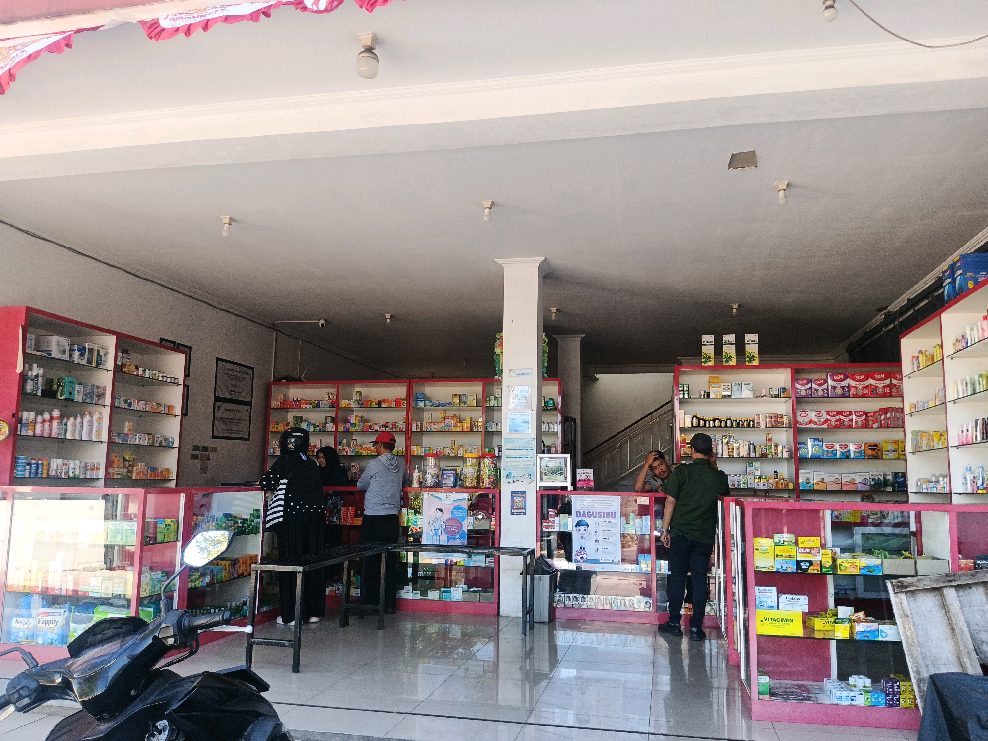 The image shows the interior of a pharmacy located in Kel. Pasir Endah, Indonesia. The pharmacy is well-stocked with shelves full of medicines, health products, and baby formula. A red and white banner with the word "INDONESIA" is visible on the ceiling, suggesting a national celebration or event. There are several individuals in the store: two women in hijabs are browsing the shelves, and two men are standing at a counter, one appearing to be a customer and the other an employee. A motorcycle is parked in the foreground, partially obscuring the view of the store. The lighting in the store is bright, coming from overhead fixtures. The floor is tiled and reflects the light. The pharmacy appears to be open for business, with customers present and products displayed for sale.