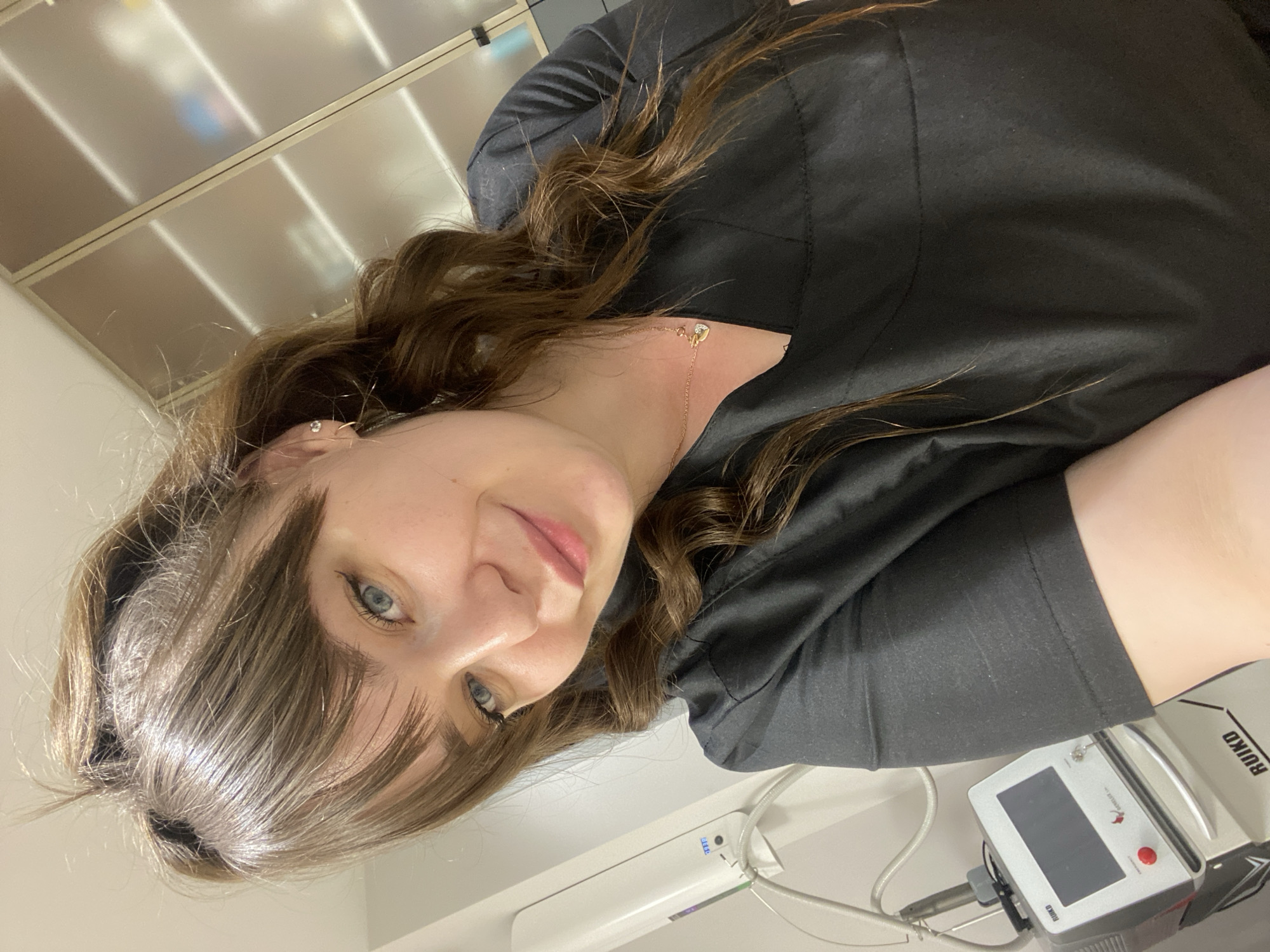 A young woman with brown hair and bangs poses in an indoor setting, likely a medical or cosmetic office. She is wearing a black top and a delicate gold necklace. To her right, a medical device labeled "RUIKD" is visible on a white surface. The background includes a cabinet with frosted glass doors and overhead lighting. There is no indication that this image was taken in Votkinsk, Russia.
