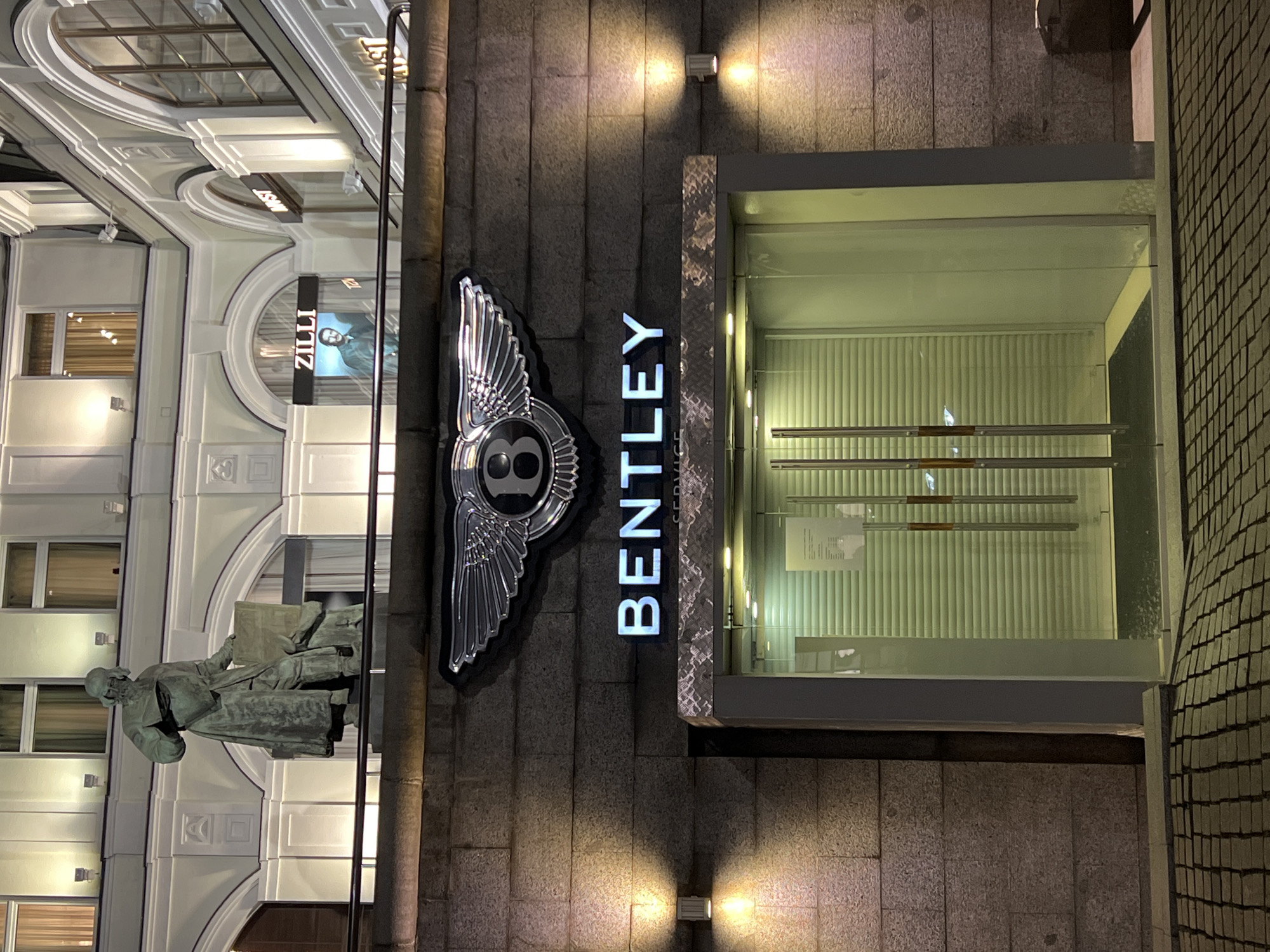 This is a medium shot, captured at night, of a Bentley service entrance. The entrance is a glass door with two large, metallic handles, set into a textured, dark grey stone facade. Above the entrance, prominently displayed, is the iconic Bentley wings emblem, rendered in chrome. Below the emblem, the word "BENTLEY" is illuminated in white neon lettering, with "SERVICE" in smaller white lettering underneath. Two wall-mounted light fixtures cast a warm glow on the stone facade on either side of the entrance. In the background, above the Bentley signage, the upper levels of a building are visible, with classical architectural details, including arched windows and decorative moldings, illuminated by warm light. A statue of a man, likely a historical figure, stands on a pedestal on an upper balcony, facing left. To the right of the statue, on a lower level, are storefronts with illuminated signage, including "ZILLI" and "MUST". The foreground shows a patterned cobblestone walkway leading towards the entrance, with a slight upward incline. The overall impression is one of upscale luxury and a formal, dimly lit urban setting. The building appears to be located in a city, and the style of architecture suggests a European, possibly Russian, location, with the "MUST" sign potentially indicating a store with international brands.