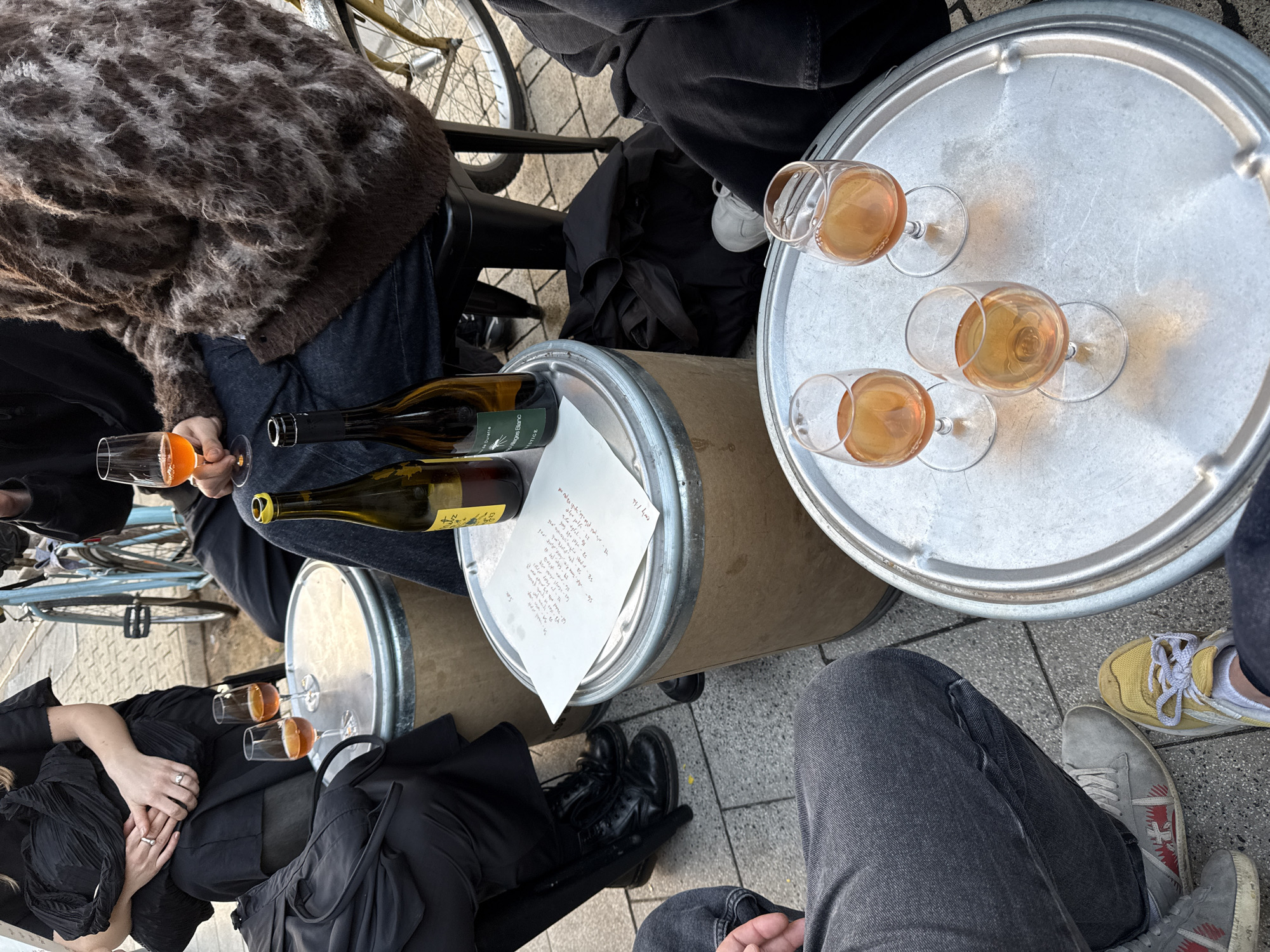 The image depicts a group of people gathered outdoors, likely in a casual urban setting, possibly Tel-Aviv, Israel. They are sitting around improvised tables constructed from what appear to be metal drums. One table is topped with a silver tray. Multiple wine glasses containing orange-colored wine are distributed on the tables. Two wine bottles are on one drum table, along with a white sheet of paper bearing handwriting. Individuals are seated around the tables, with various fashion choices, including black clothing, patterned knitwear, jeans, and an array of footwear ranging from sneakers to boots. A bicycle is partially visible in the background.