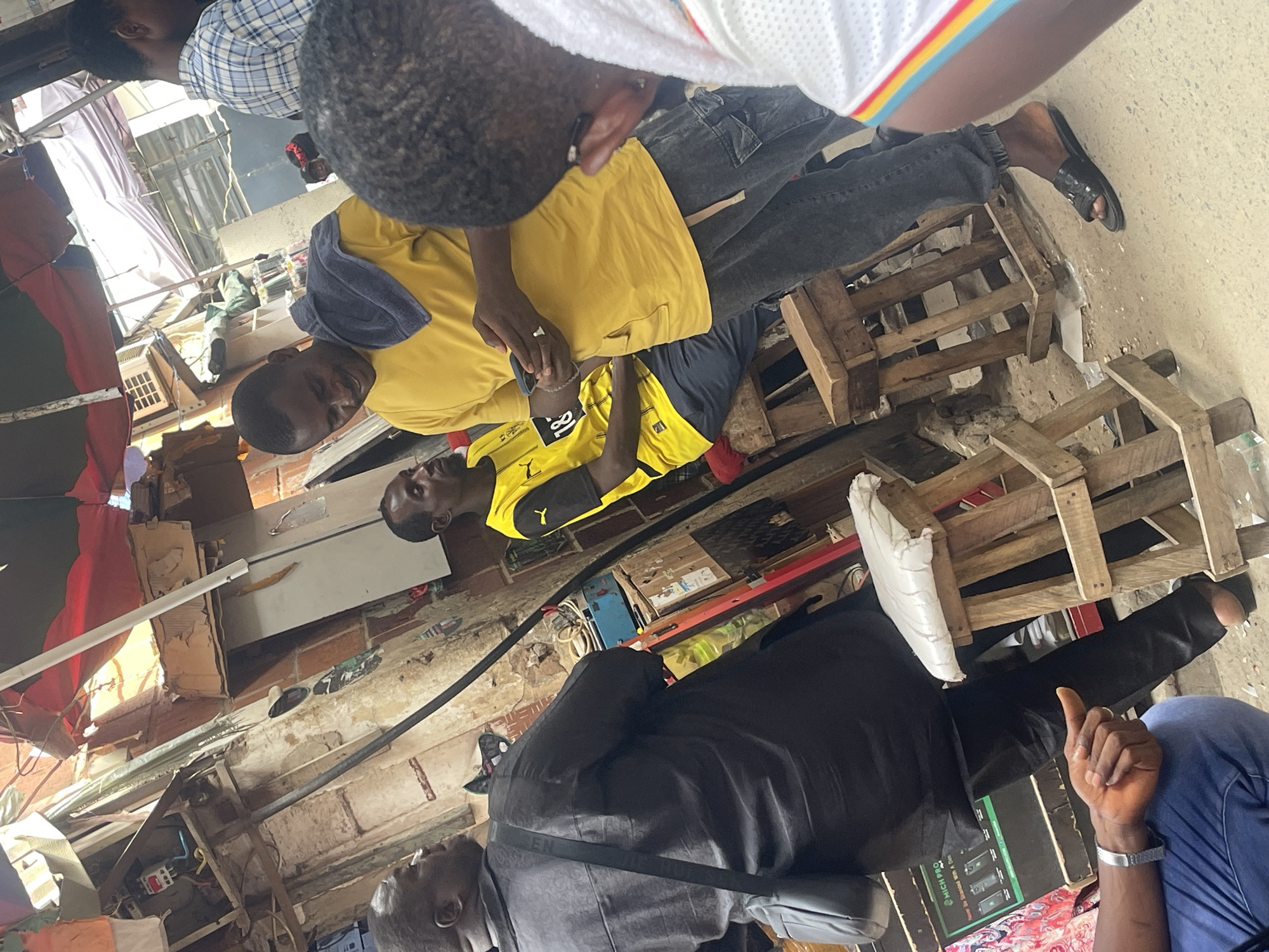 The photograph depicts an outdoor scene likely in a crowded market area of Abuja, Nigeria. Several individuals are gathered, some wearing casual clothing like a yellow t-shirt and jeans, while another is dressed in a black suit. One man is wearing sandals. They appear to be engaged in a discussion or transaction. There are wooden pallets stacked on the ground, suggesting a marketplace or informal business setting. The background shows a somewhat cluttered environment with various buildings, electrical installations, and materials like cardboard boxes, indicative of commercial activity.