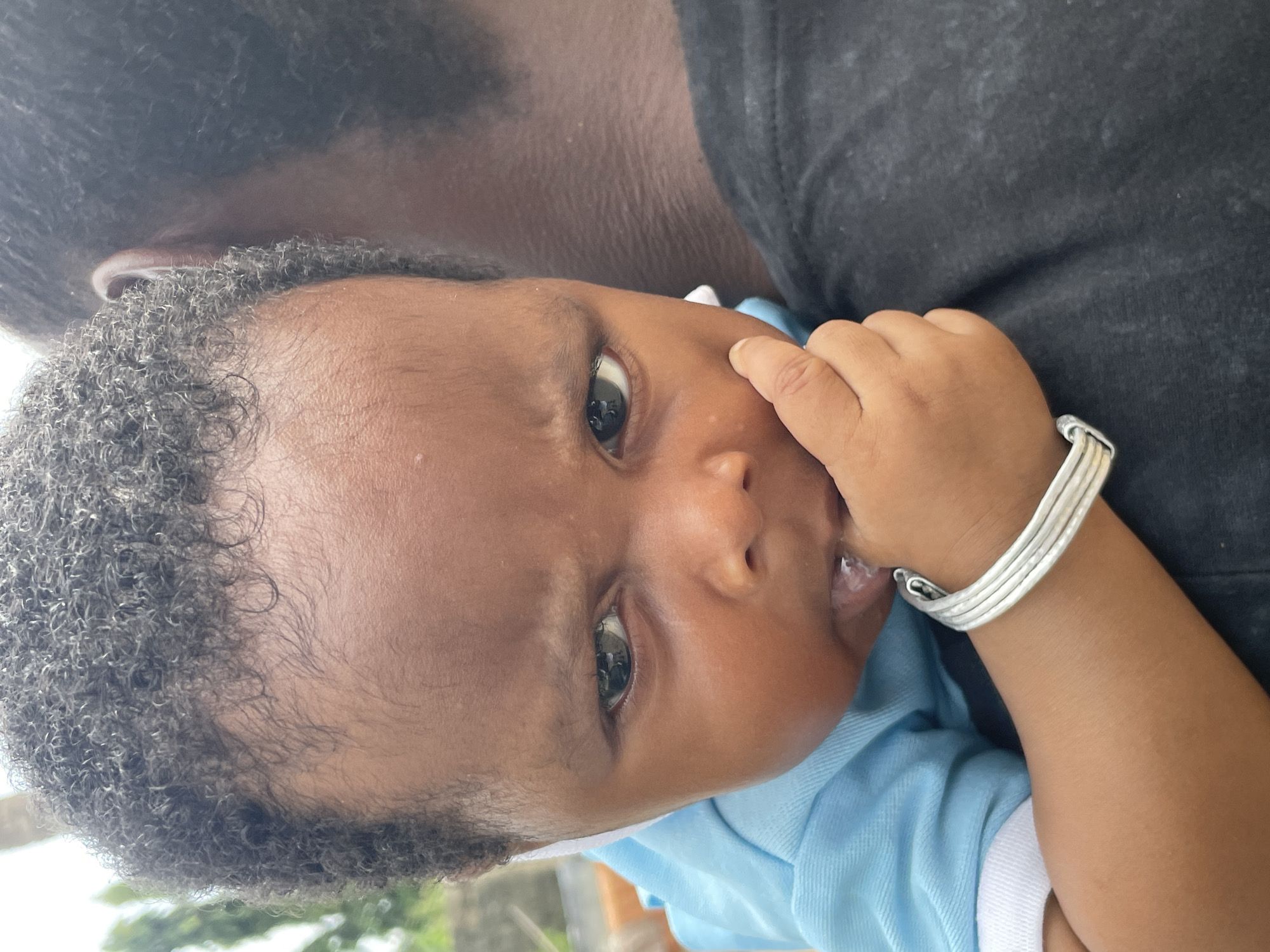 A baby is being held, with the baby's face clearly visible. The baby has dark skin, curly black hair, and is wearing a light blue shirt. The baby has a silver bracelet on its wrist. The baby's fingers are near its mouth. A person with dark skin and wearing a black shirt is holding the baby. The background is slightly out of focus, but greenery is visible, suggesting an outdoor location. No details suggest the location is Badagry, Nigeria.