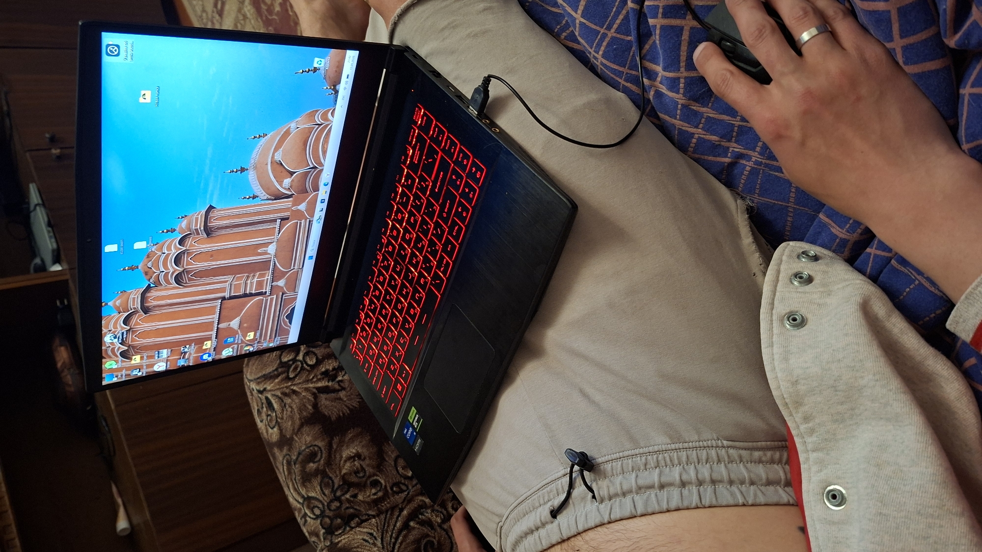 A person is sitting indoors with a laptop resting on their lap. The laptop screen displays an image of a large building under a blue sky. The laptop's keyboard is illuminated with red light. A computer mouse is visible in the person's right hand, which has a ring on one of the fingers. The person is wearing light-colored casual clothing. Part of a patterned fabric is visible underneath the person. The immediate environment suggests a casual, relaxed setting, possibly in a home or a private space. There is no information in the image about the location being in the city of Забалоцкі сельскі Савет, Belarus.