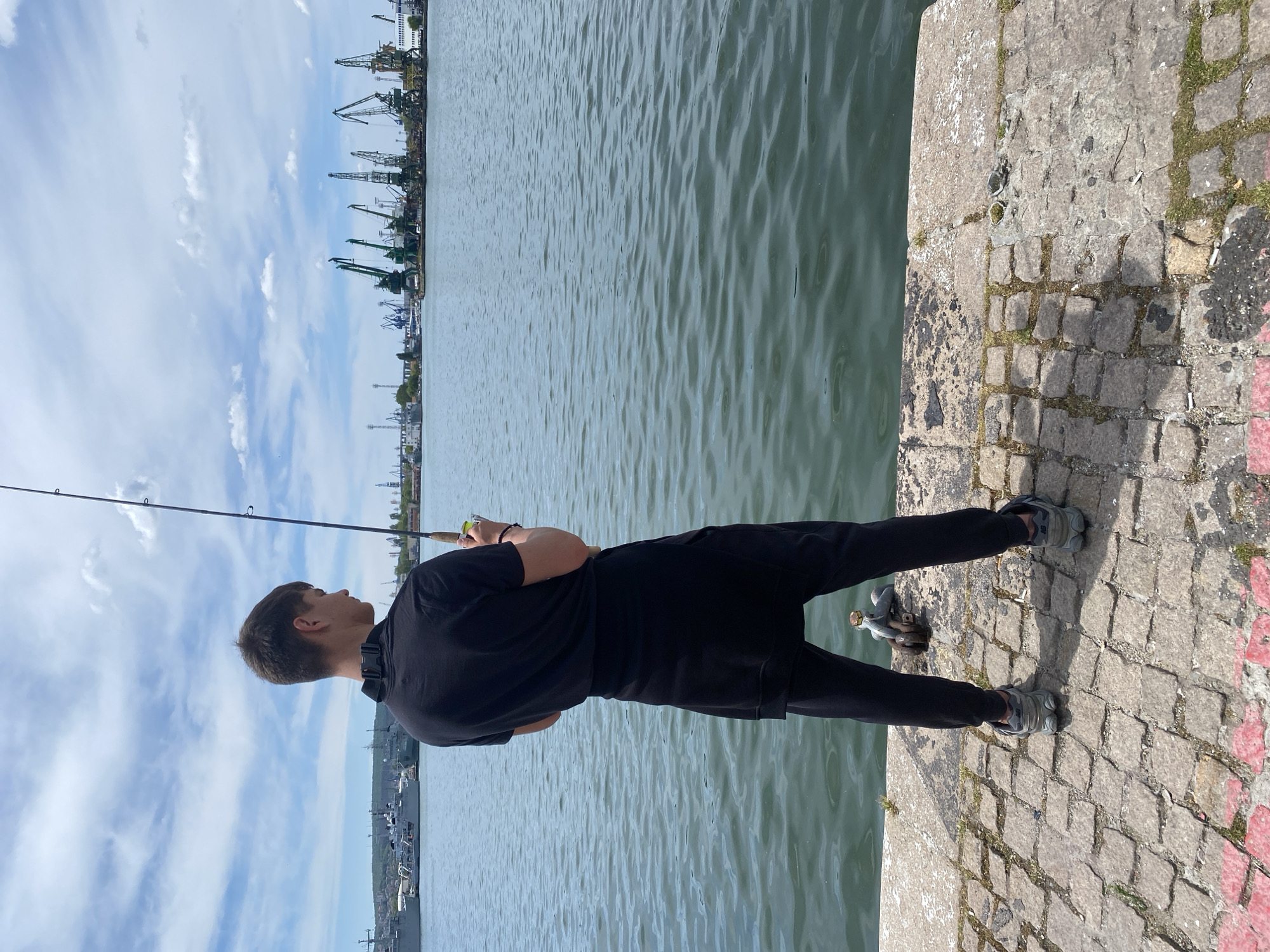 A young man in black clothing is fishing off a stone embankment. He stands with his feet parallel to the edge of the embankment, facing the water. He holds a fishing rod. The water appears calm. In the distance are industrial cranes. The embankment consists of small, rough stone blocks. The scene is bathed in daylight. The sky is partly cloudy. Given the presence of industrial cranes and the overall setting, the location is likely along the Port of Varna in Varna, Bulgaria.