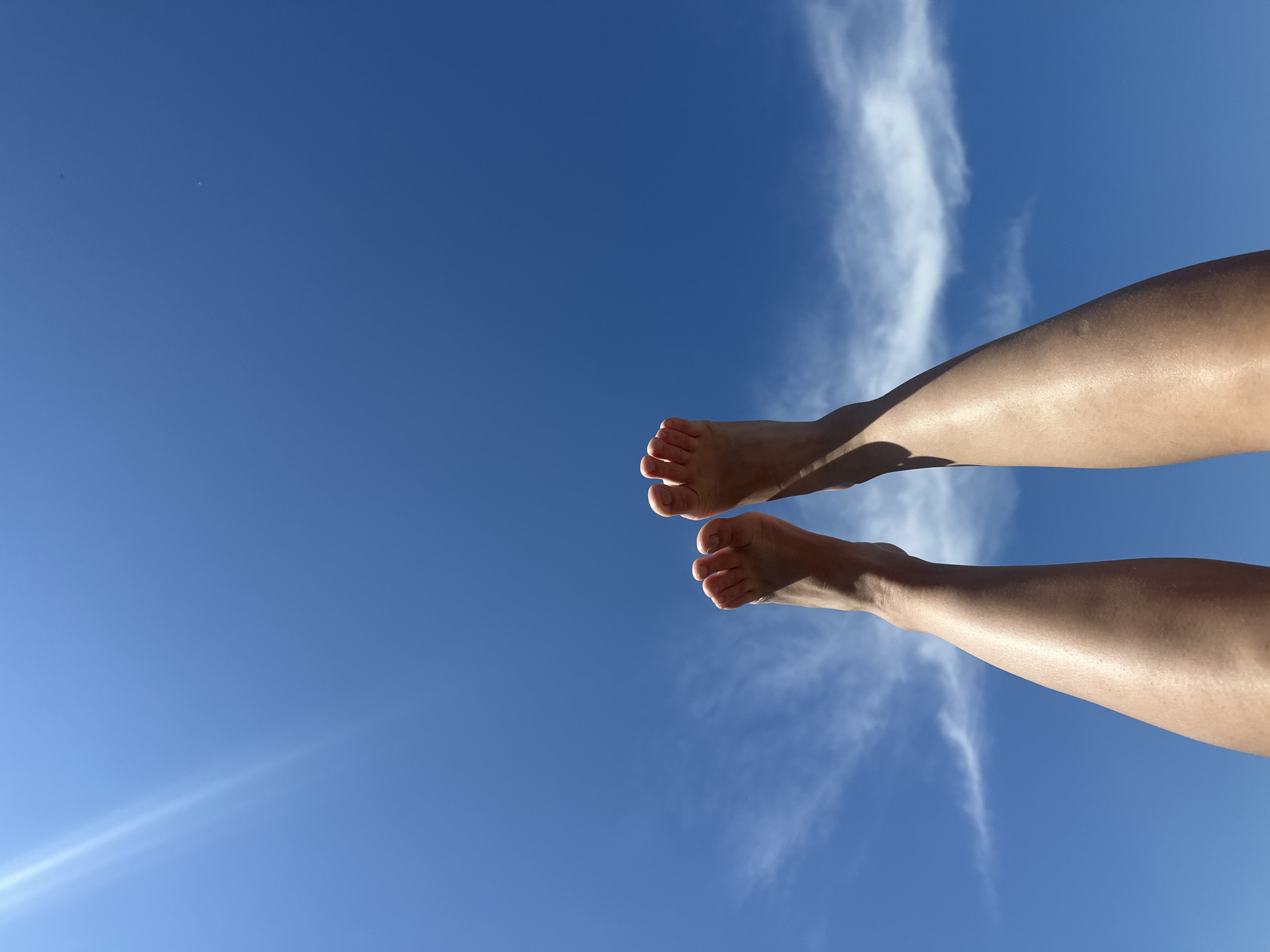 In Bely Yar, Russia, a person's bare feet and lower legs are raised against a clear blue sky with scattered wispy clouds. The sun is shining brightly, creating a lens flare in the upper left corner of the image. The skin on the legs appears tanned and is catching the sunlight. The feet are positioned in the lower half of the frame, with toes visible.