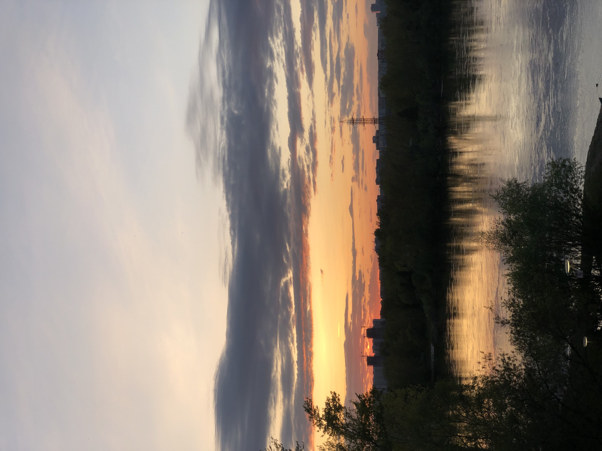 The image captures a sunset scene over a body of water. The sky features a mix of light and dark clouds, with the setting sun casting orange and yellow hues across the horizon. A silhouette of buildings and an electricity pylon are visible along the shoreline. The water reflects the colors of the sky. Trees line the foreground, partly obscuring the view. Based on the presence of the Yenisey River and its urban landscape, this could be a sunset view in Krasnoyarsk, Russia.