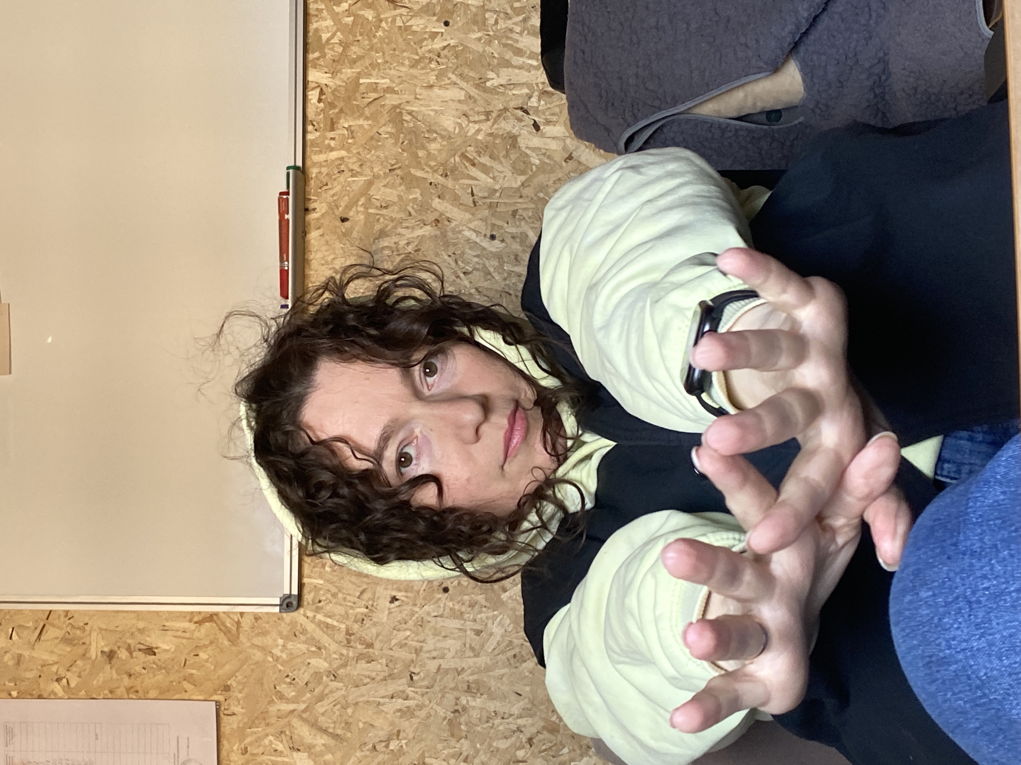 A woman with curly brown hair is seated in front of a wall made of OSB (Oriented Strand Board). She is wearing a black and pale yellow hooded jacket and jeans. She is gesturing with both hands towards the camera, with the fingers spread apart. There is a whiteboard on the left side of the image, attached to the wall, with a red marker visible. A gray garment is draped nearby. The location in the city of Ol'khovskoye, Russia is not discernible from the image.