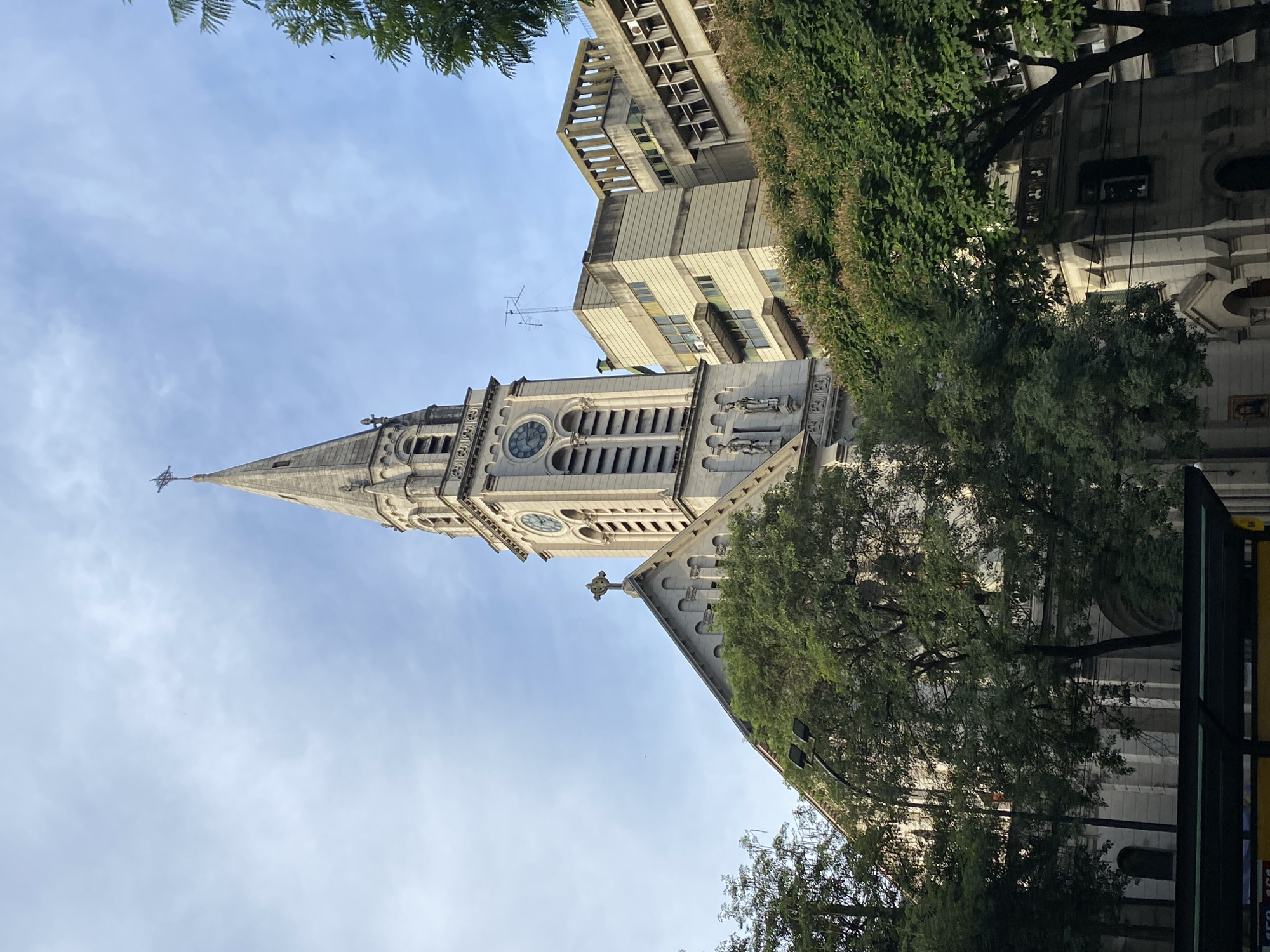 The image depicts a view of the steeple and clock tower of the Basilica del Santísimo Sacramento in Buenos Aires, Argentina, from a low angle. The architecture of the church is visible with its pointed spire, clock face, and Gothic-style detailing. Adjacent to the church, an office building with multiple levels and protruding balconies can be seen. The building is a pale beige color with dark-framed windows. Trees with green foliage partially obscure the church and building. The sky above is pale blue with scattered clouds.